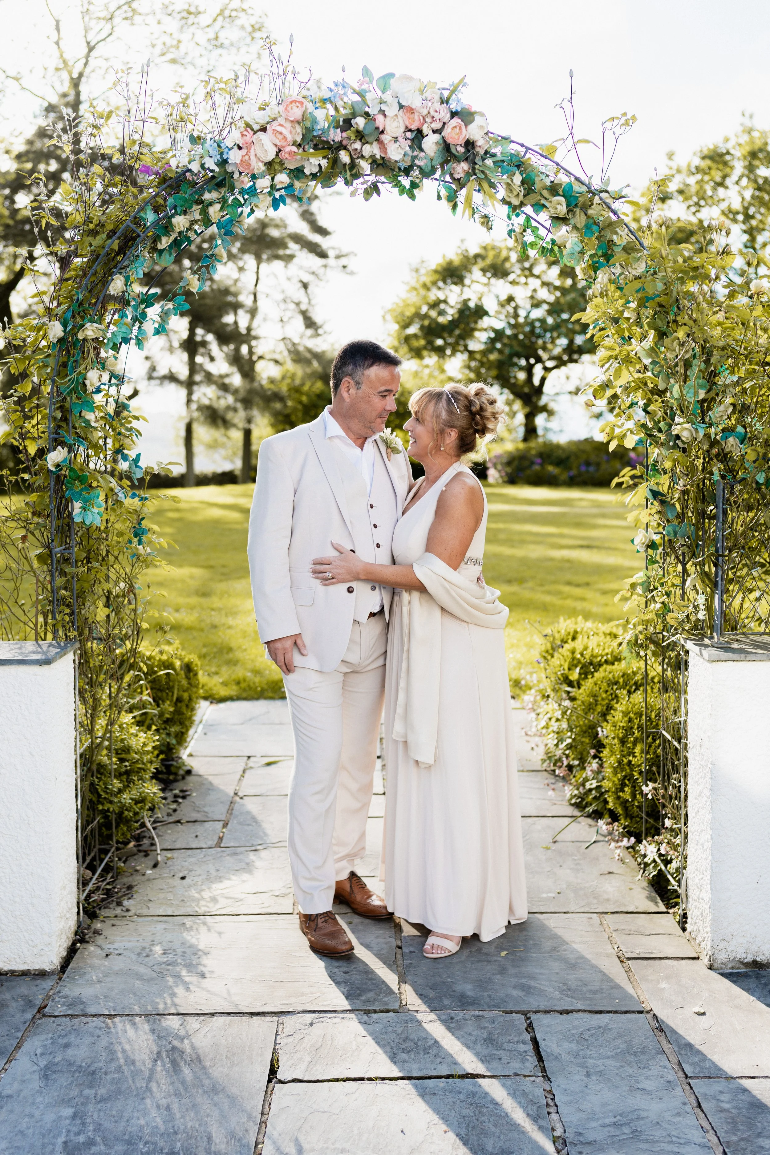 A couple in wedding attire standing under a floral arch outdoors on a sunny day.