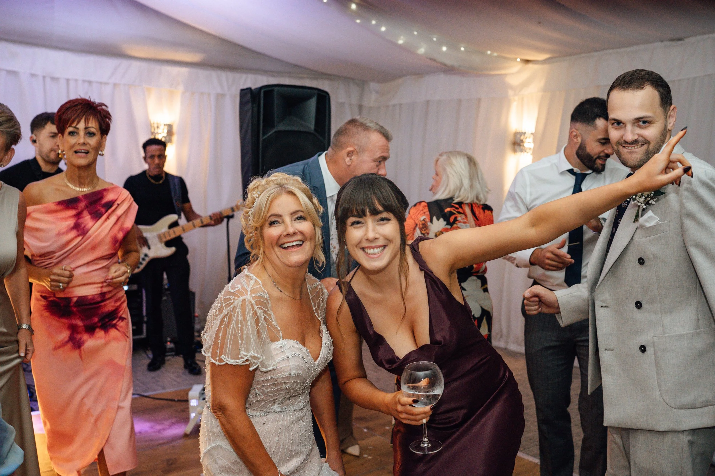 People dancing and celebrating at a wedding reception in a decorated tent, with a live band playing in the background.