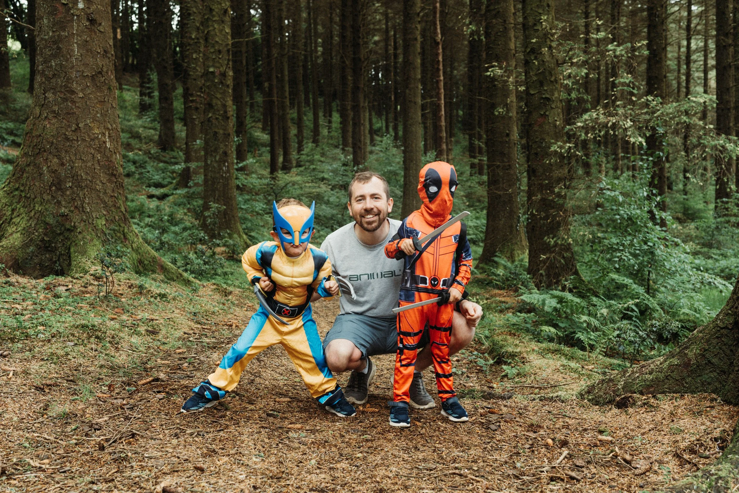 A man crouching in a forest surrounded by two children dressed as superheroes, one in a yellow and blue costume with a mask, and the other in an orange and black costume with a mask and swords.