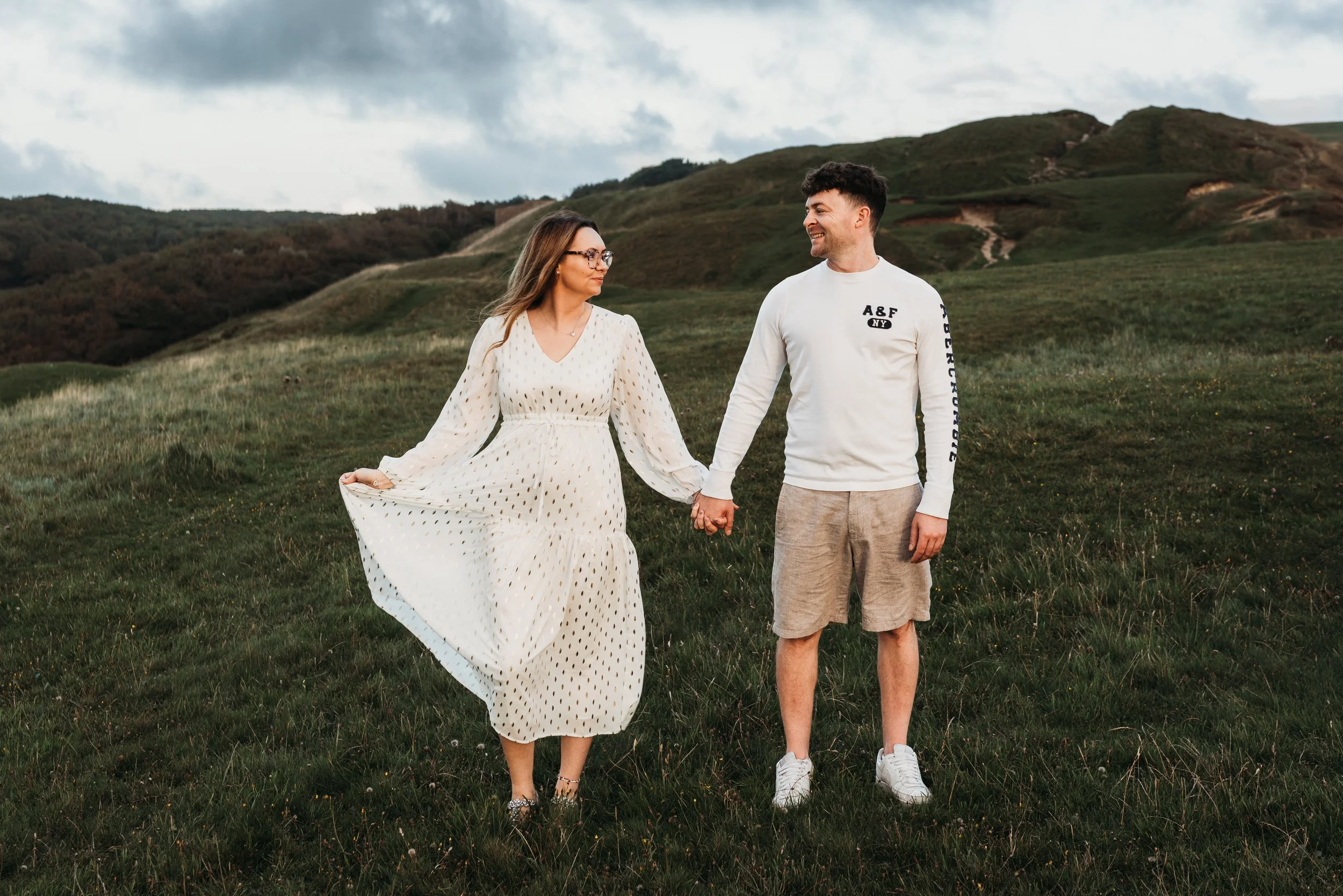 A young woman in a white dress holding hands with a young man in casual clothes, walking through a grassy field with green hills in the background under a cloudy sky.