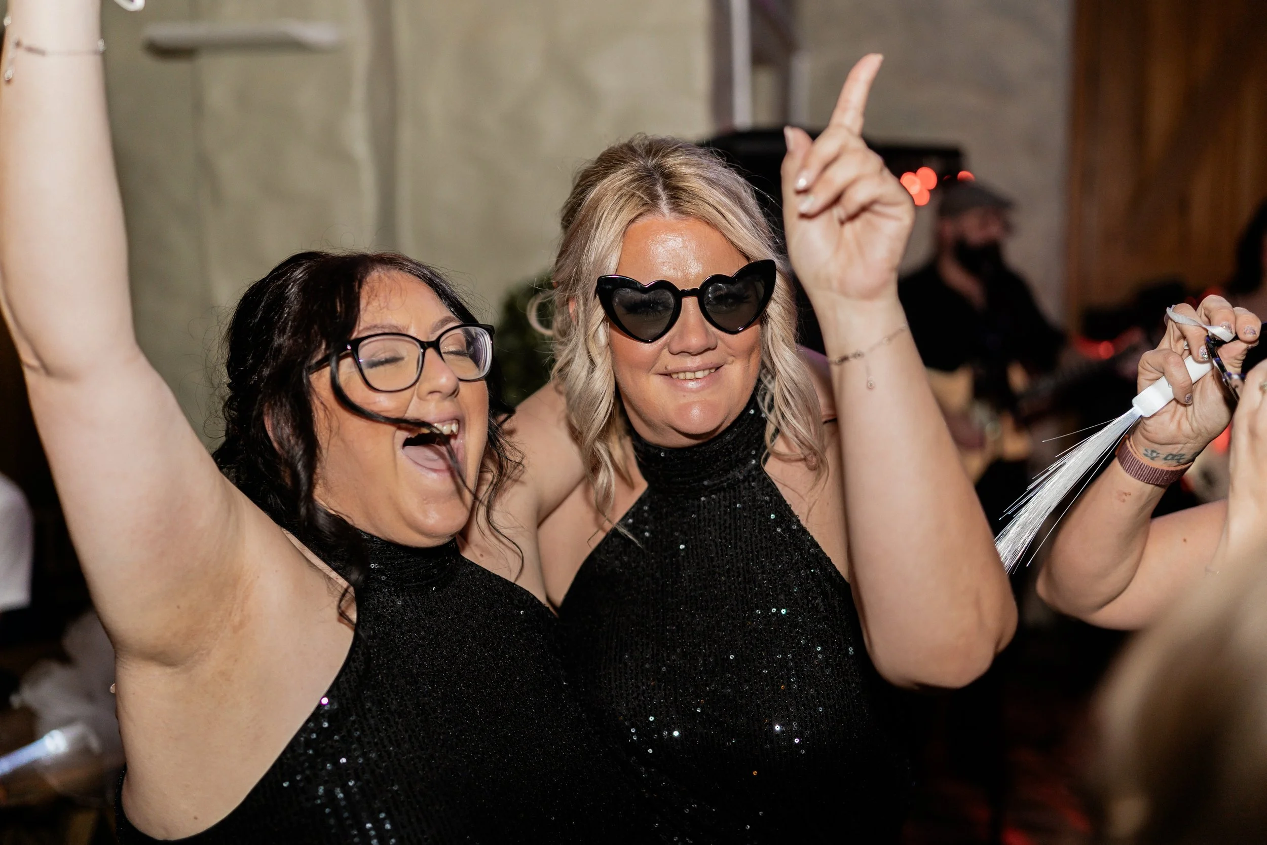 Two women wearing black sequined dresses and sunglasses, dancing and celebrating at a party.