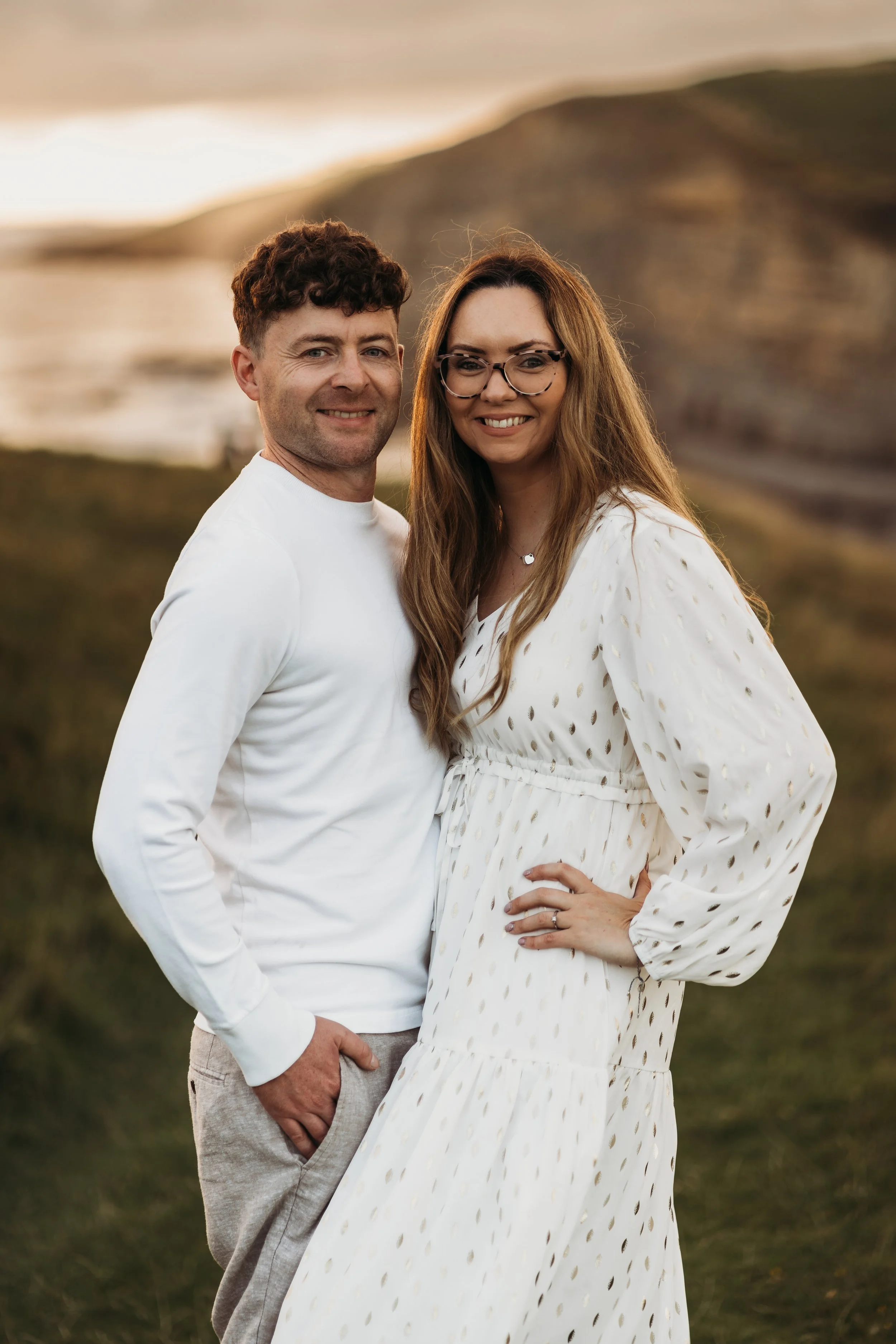 A smiling couple outdoors, standing on grass with a hilly landscape and water in the background, during sunset.