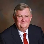 Smiling man with gray hair wearing a dark suit jacket, white shirt and red tie.