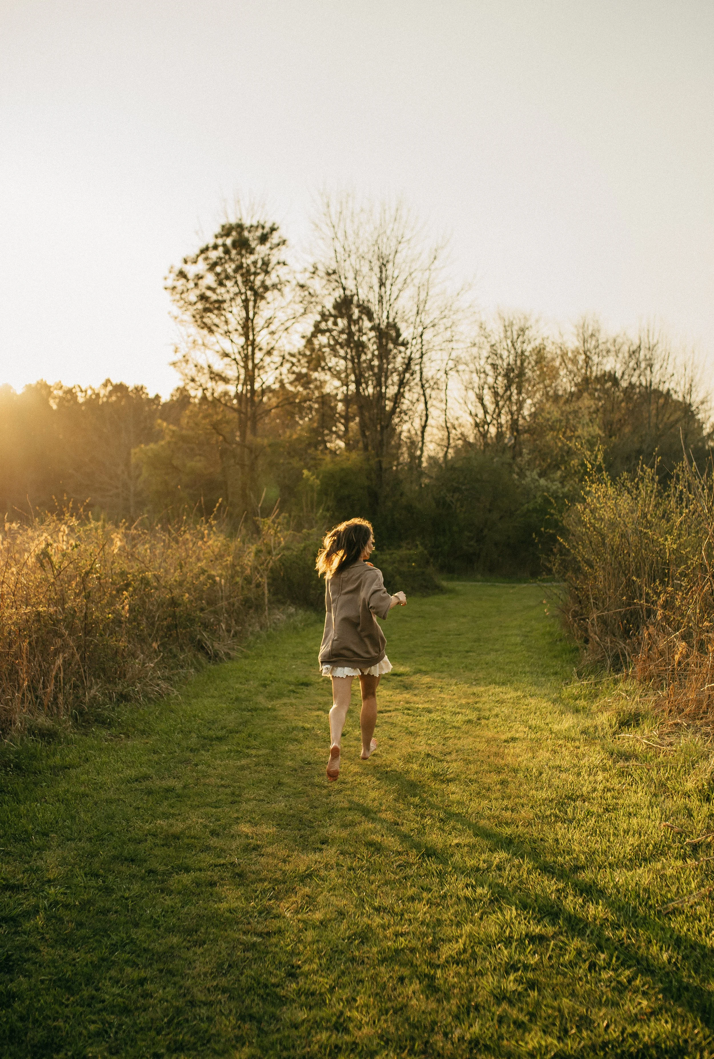 A girl running along a grassy path in a park during sunset, surrounded by trees and bushes.