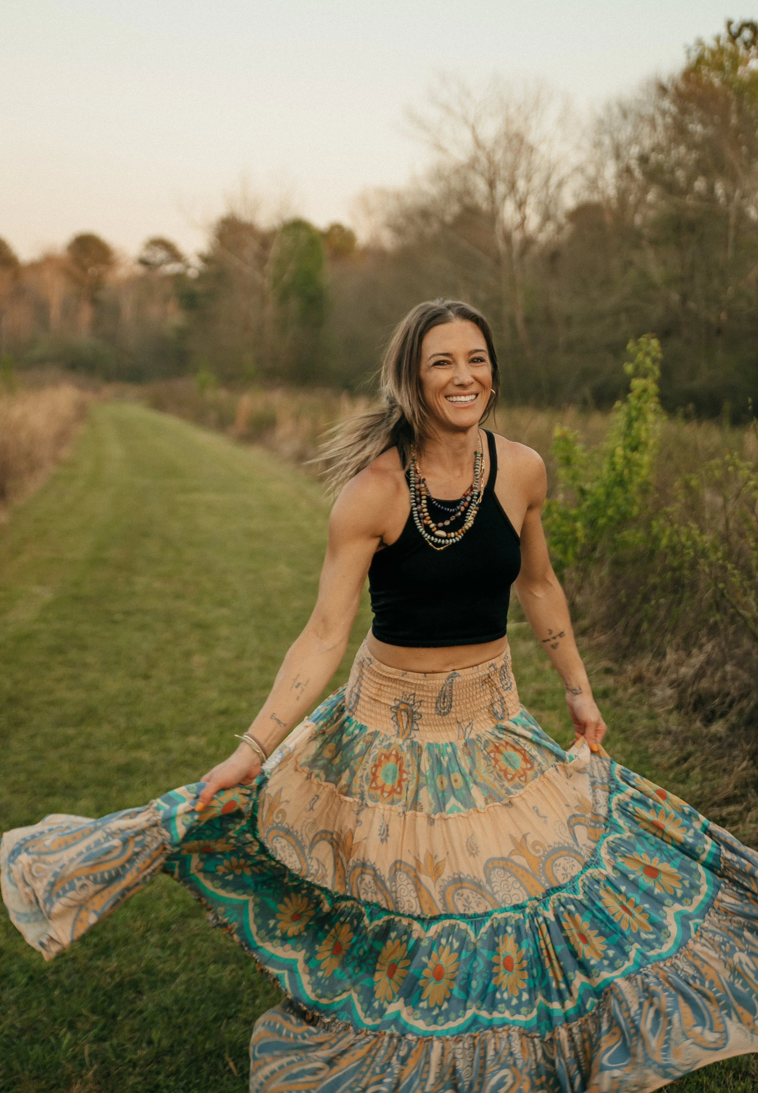 A woman in a black tank top and colorful bohemian skirt smiling and spinning outdoors on a grassy path with trees in the background.