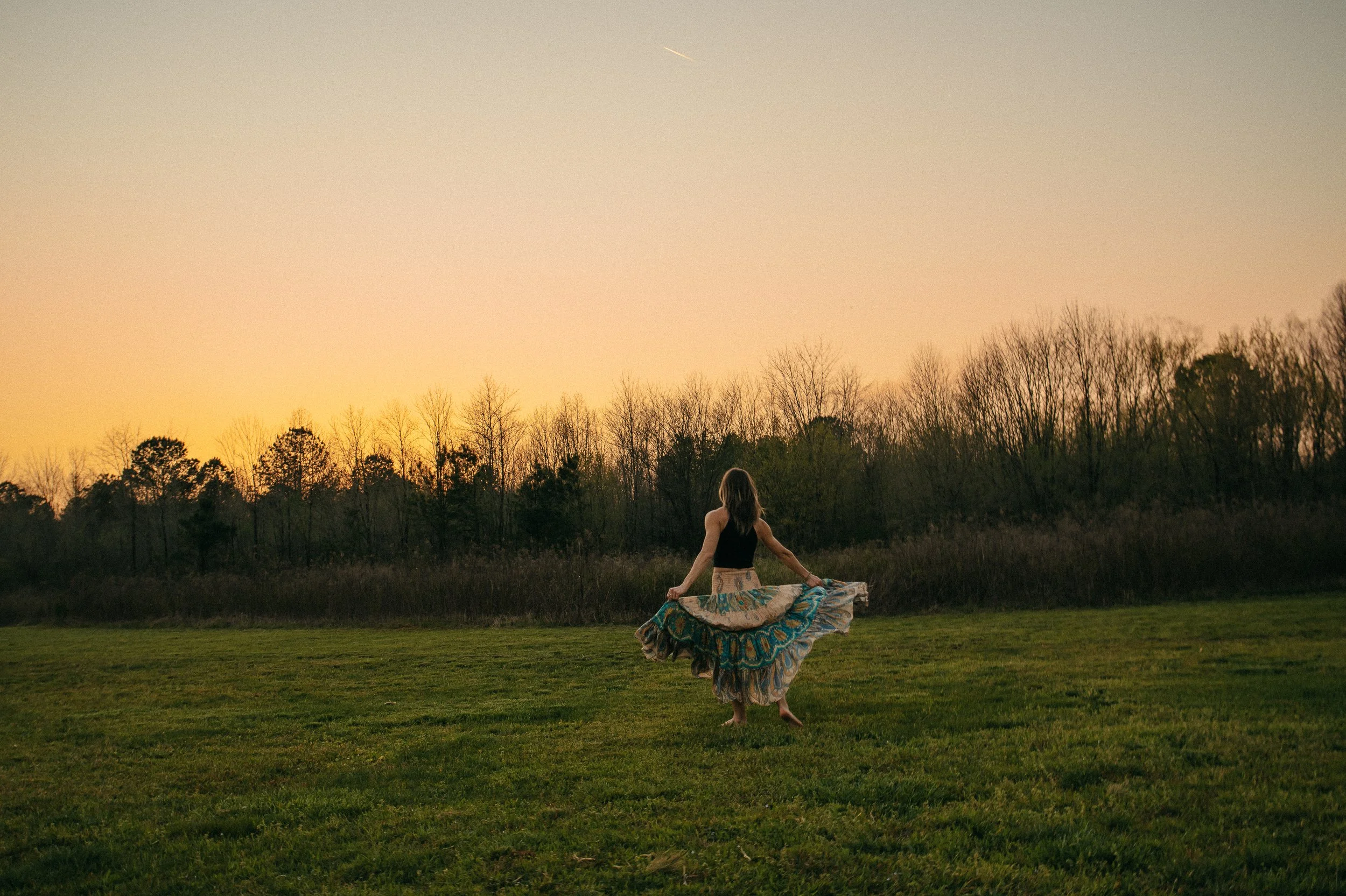 A woman with long hair is dancing on a grassy field during sunset, wearing a black top and a colorful, flowing skirt.