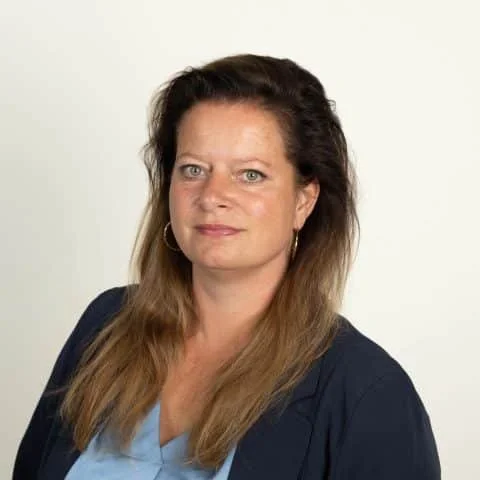 Portrait of a woman with long brown hair, wearing a navy blazer and a light blue shirt, standing against a plain white background.