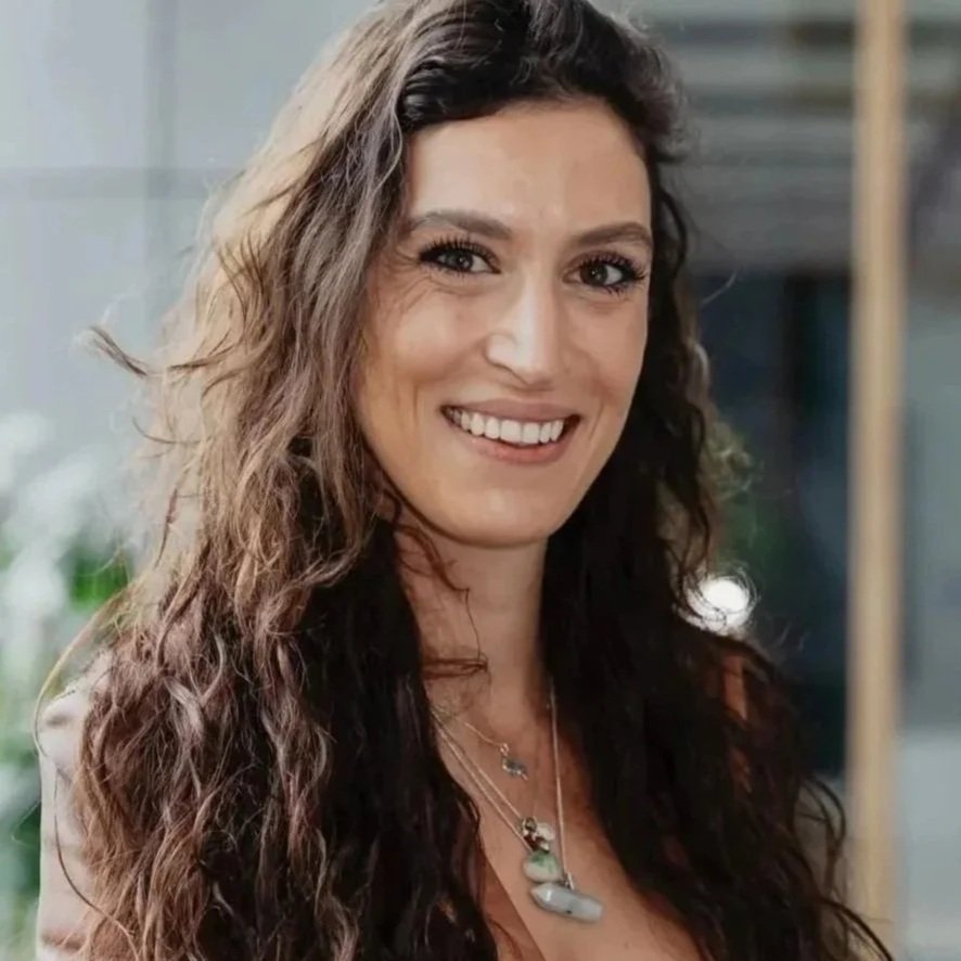 A woman with long, curly brown hair smiling at the camera. She is wearing a necklace with green stones and a beige top. The background is blurred with some indoor elements.
