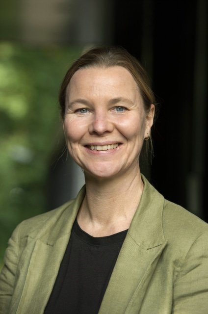 A woman with light skin, brown hair, and blue eyes smiling, wearing a black top and a green blazer, standing outdoors with blurred greenery in the background.