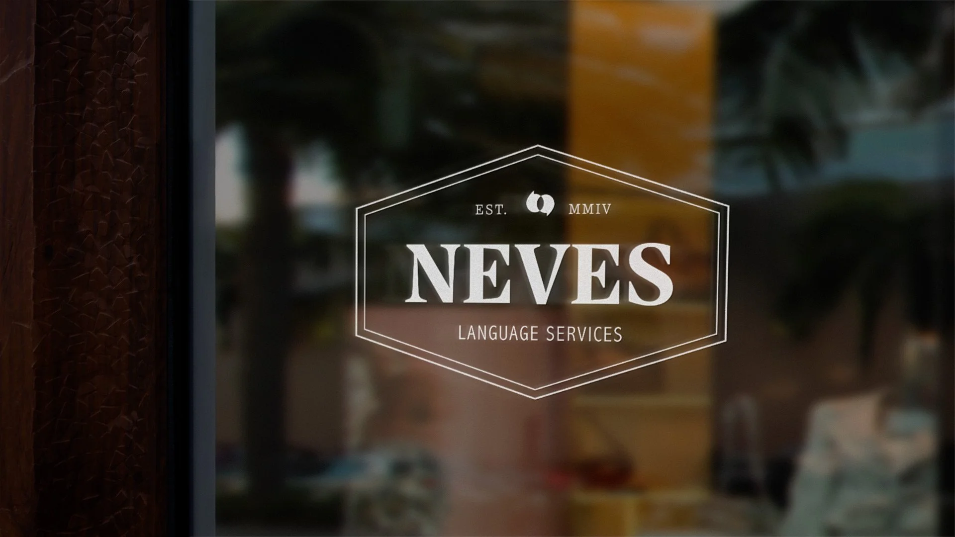 A glass door with a white logo and text that reads 'NÉVES LANGUAGE SERVICES', established in MMIV, with a background of blurred outdoor scene.