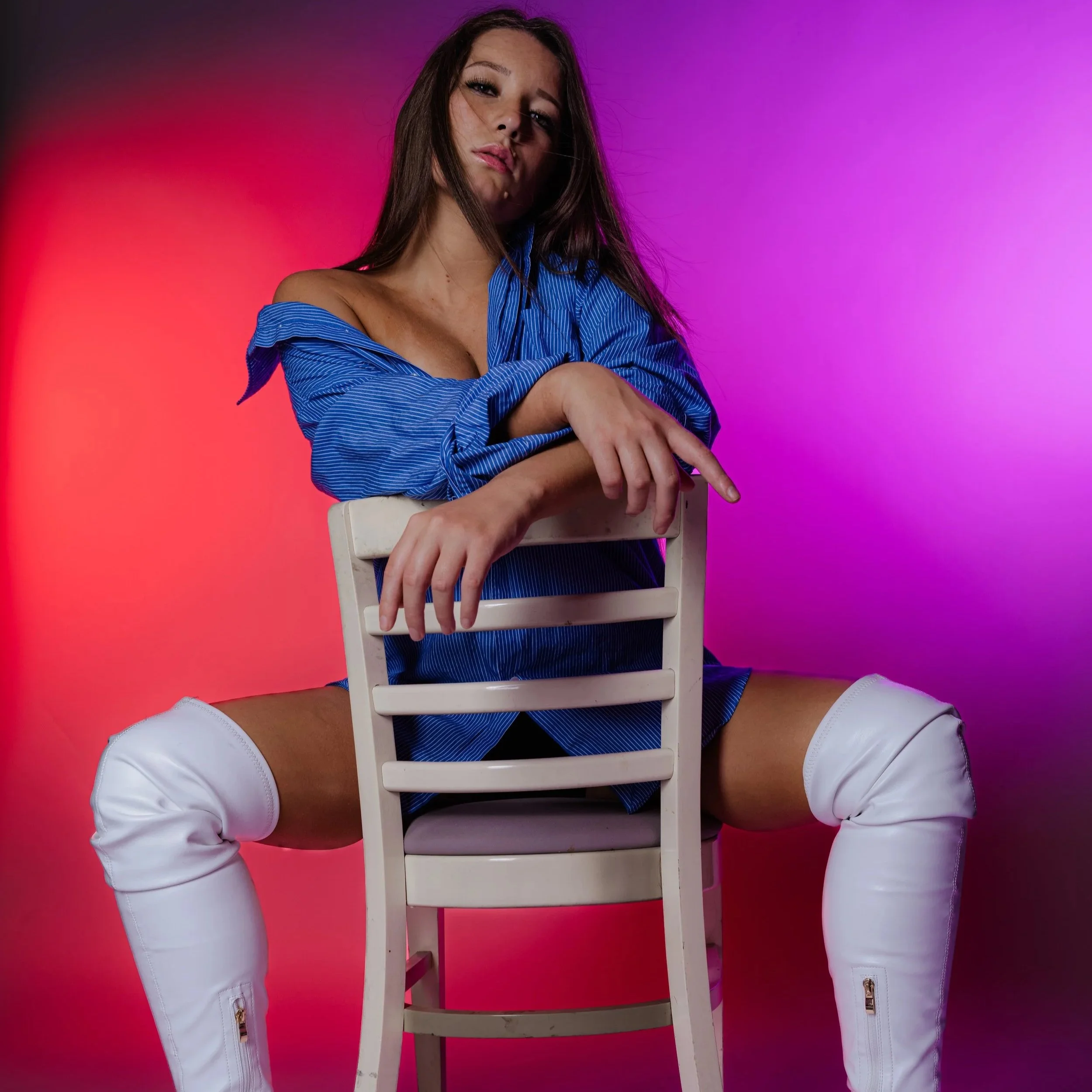 A woman in a blue shirt and white thigh-high boots sitting on a white chair, with pink and purple background lighting.