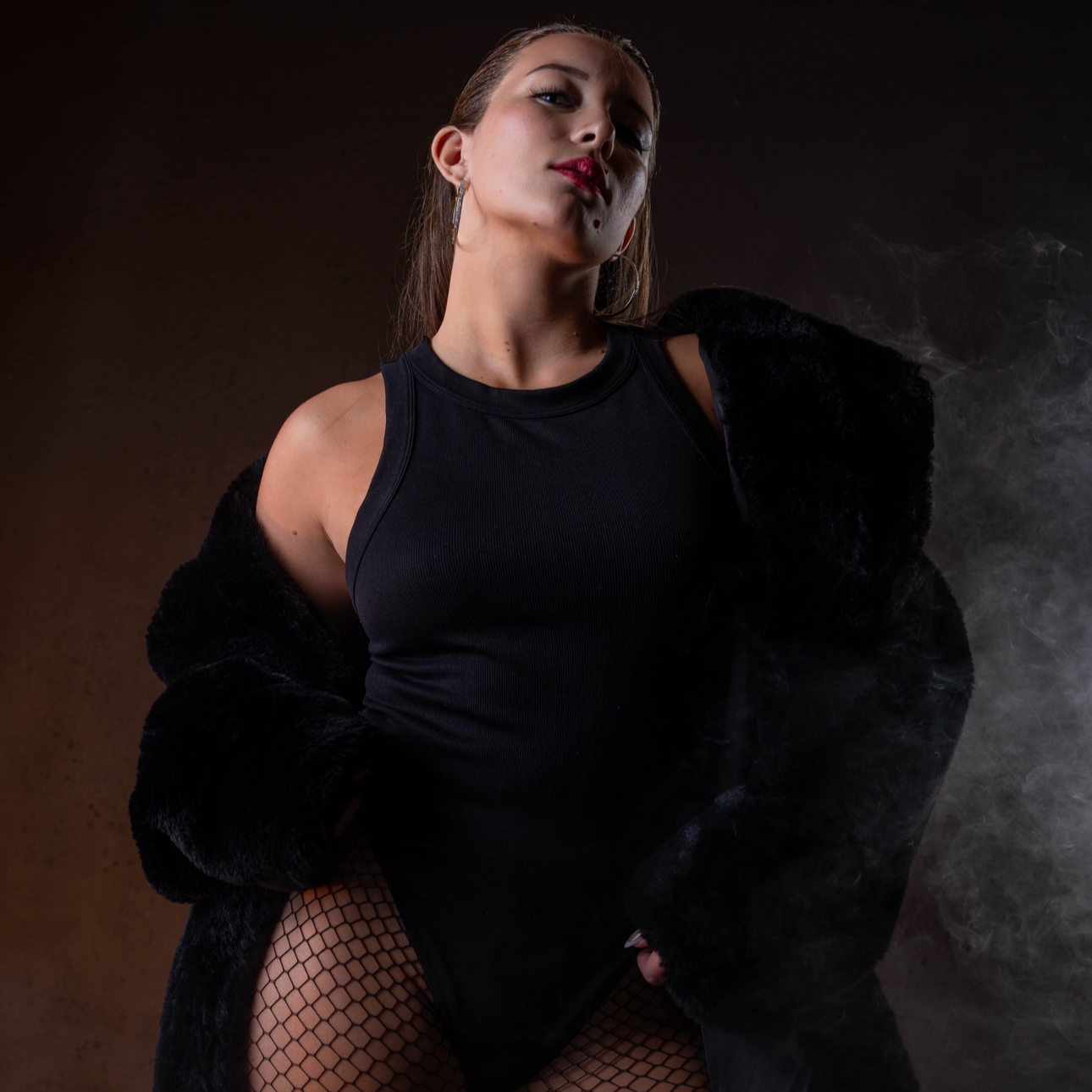A woman poses confidently in a black sleeveless top, black shorts, fishnet stockings, and a black fur coat in a dark, smoky setting.