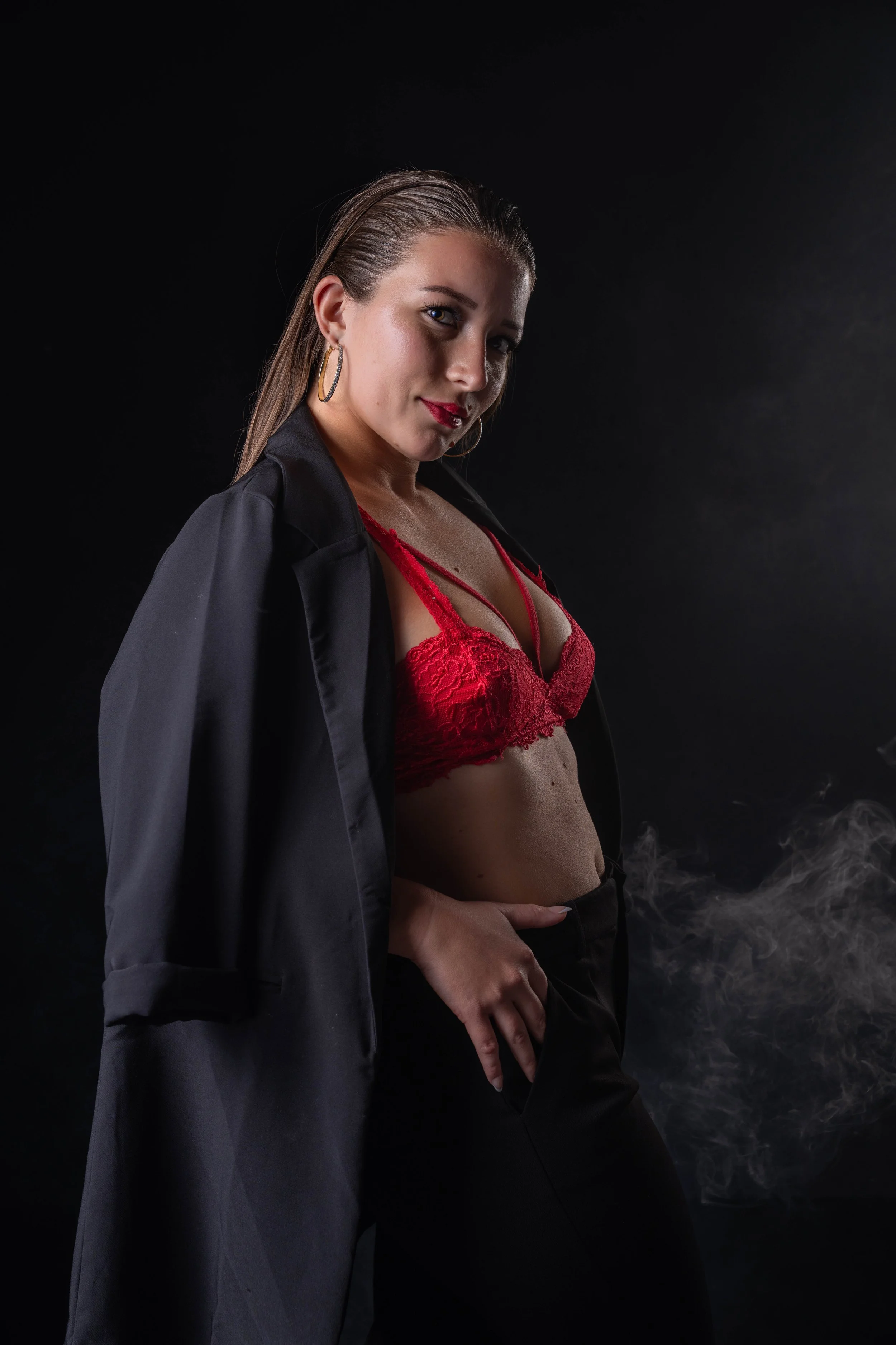 A woman wearing a black jacket off her shoulders, a red lace bra, and black pants, posing against a dark background with some smoke or mist.