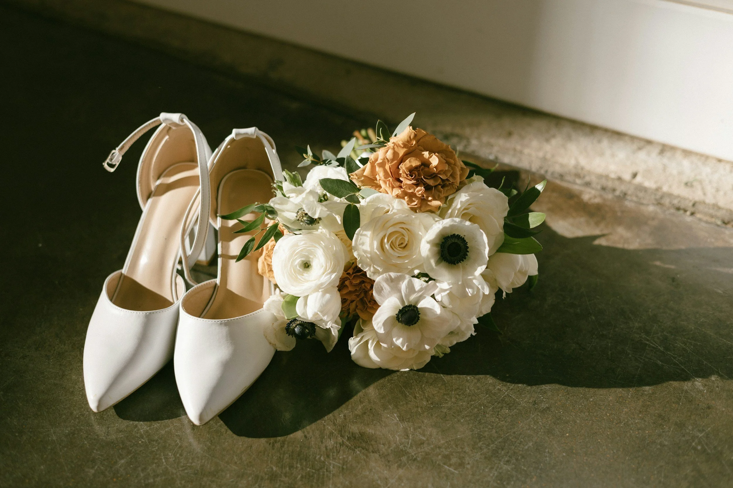 A pair of white high-heeled shoes placed next to a bouquet of white and beige flowers on a dark surface.