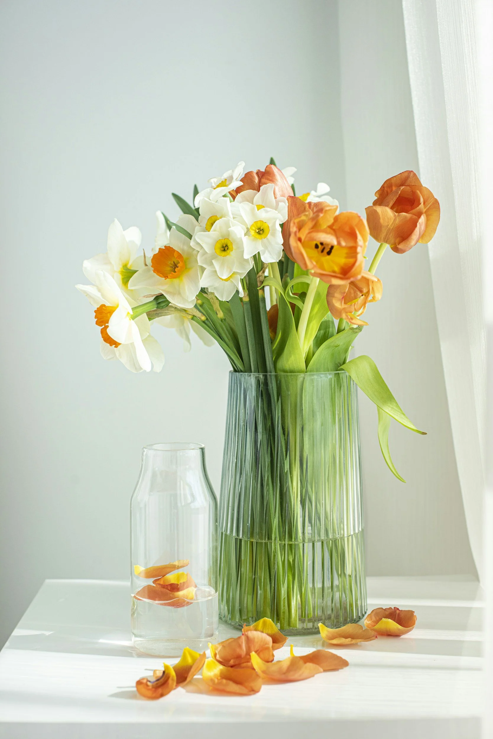 A clear glass vase with fresh orange and white flowers, including daffodils and tulips, on a white surface by a window with sunlight.