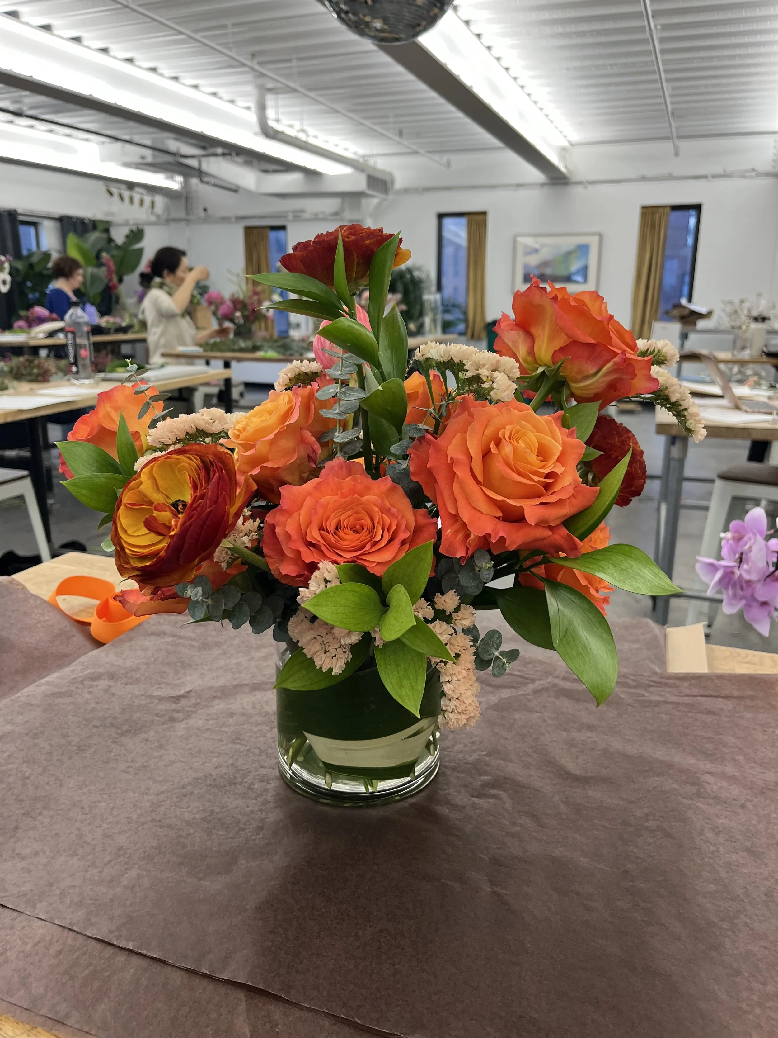 A colorful flower arrangement with orange roses, red and yellow ranunculus, light pink carnations, green foliage, in a glass vase on a table. In the background, people are working on flower arrangements in a bright, industrial-style room.