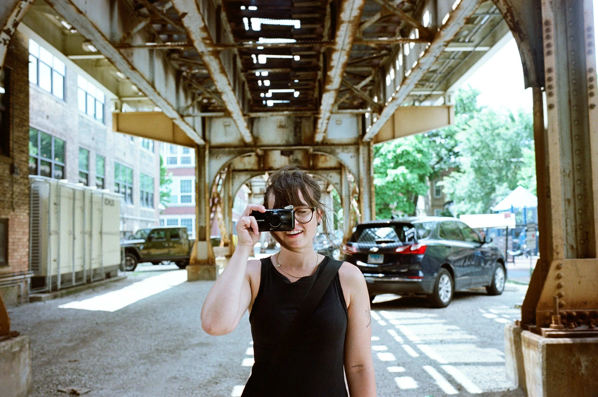 A smiling young woman with glasses taking a photo with a camera under a rusty steel bridge in an urban setting on a sunny day.