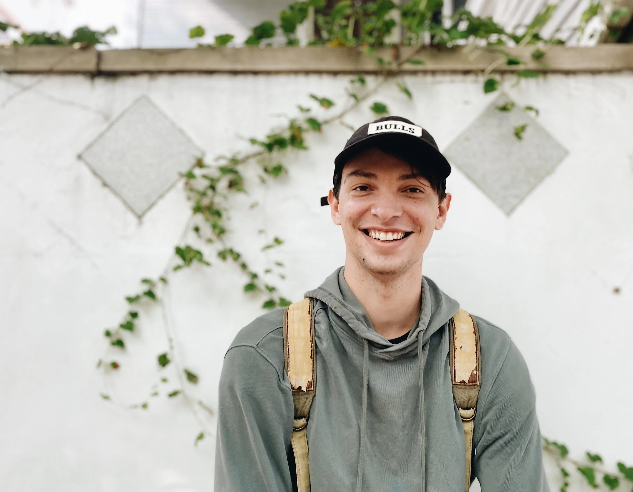 A young man smiling, wearing a black cap with the word "BULLS" on it, a gray hoodie, and backpack straps, standing outdoors in front of a white wall with diamond-shaped concrete blocks and green vines.