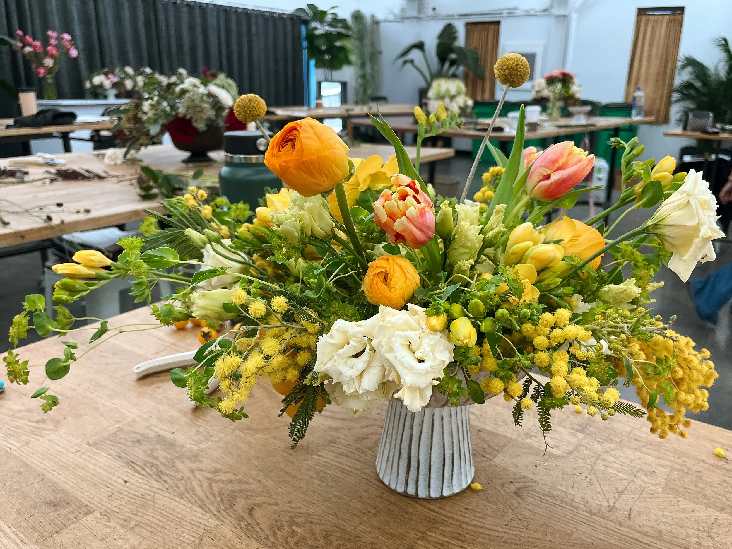 A large floral arrangement with yellow, white, orange, and pink flowers in a white vase on a wooden table in a room with tables, chairs, and plants.