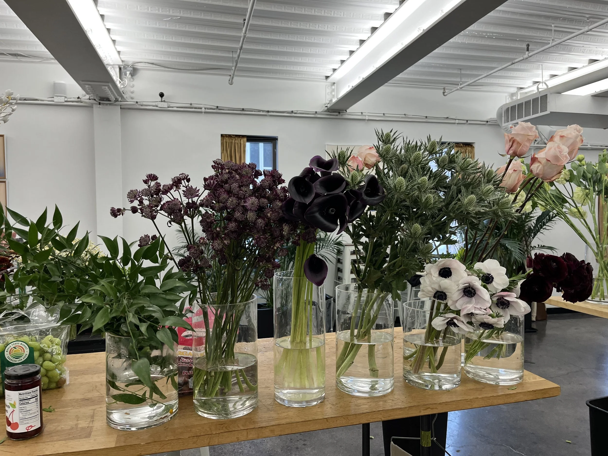 Various cut flowers in glass vases on a wooden table inside a room with white walls and a high ceiling.