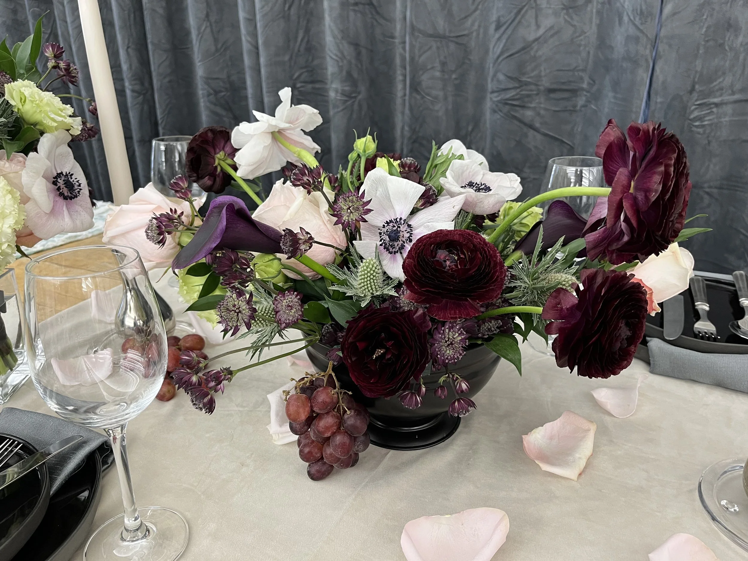 A floral centerpiece with dark red, white, and purple flowers, arranged in a black vase on a table set with wine glasses, silverware, and pink rose petals scattered on the tablecloth.