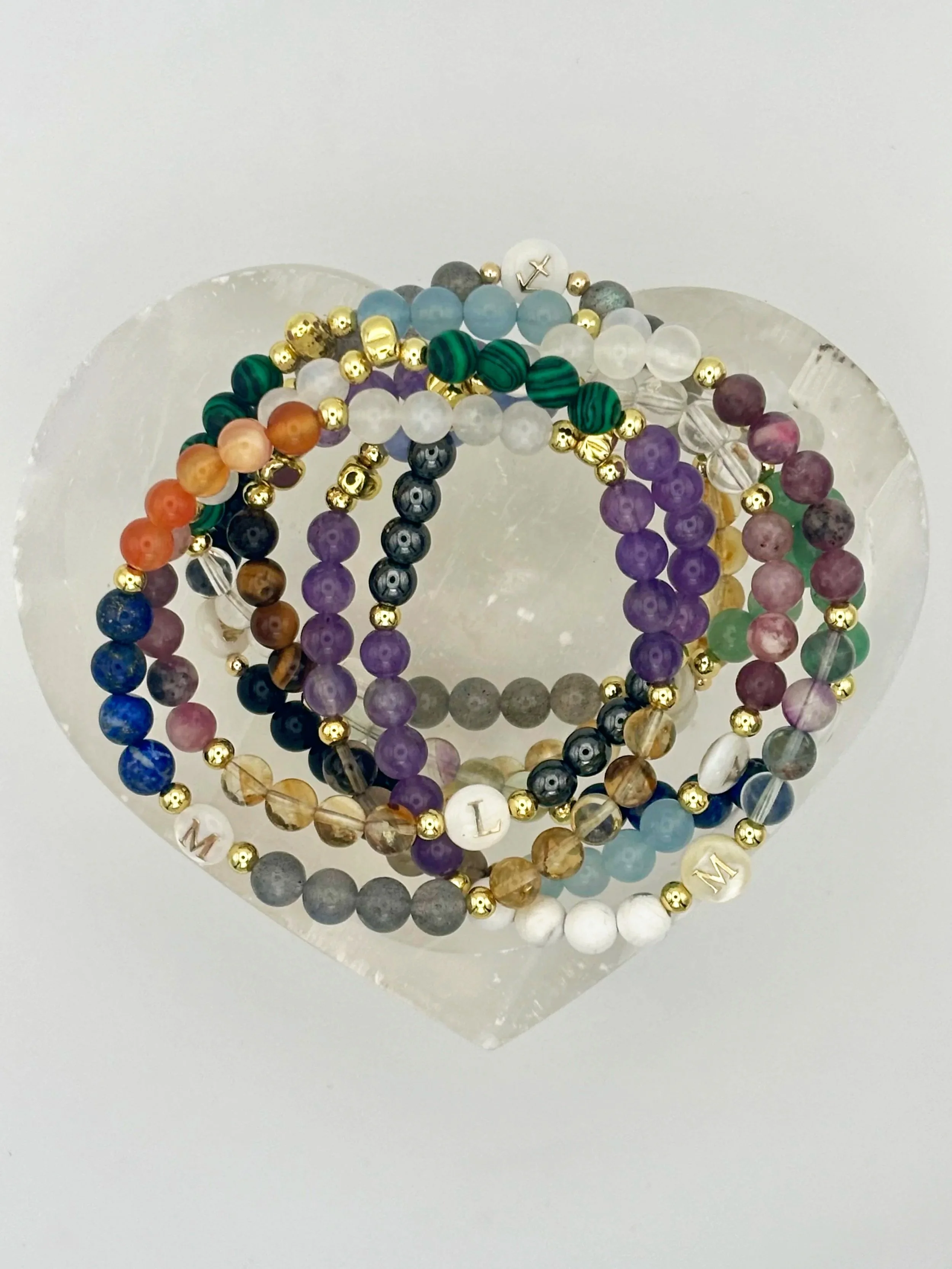 Display of multiple beaded bracelets with colorful beads, including purple, green, white, black, and gold, arranged on a clear, heart-shaped stand against a plain white background.