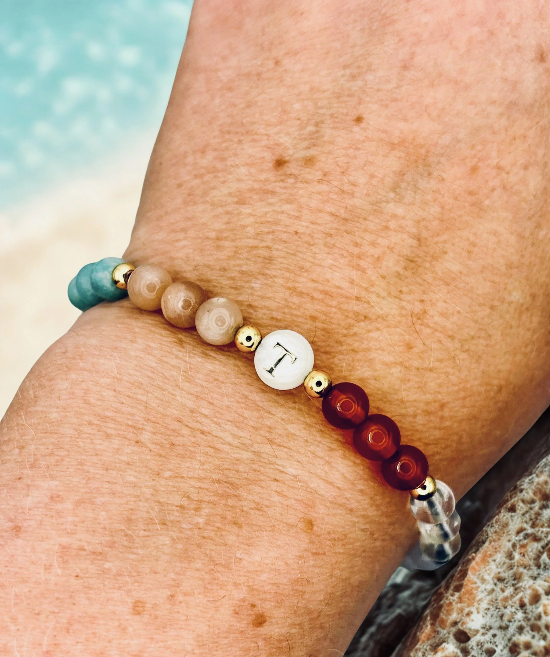 Close-up of a person's skin with a sunspell crystal bracelet on their wrist. The skin has some freckles and fine hair, and the background appears to be a blurred beach scene with sand and water.