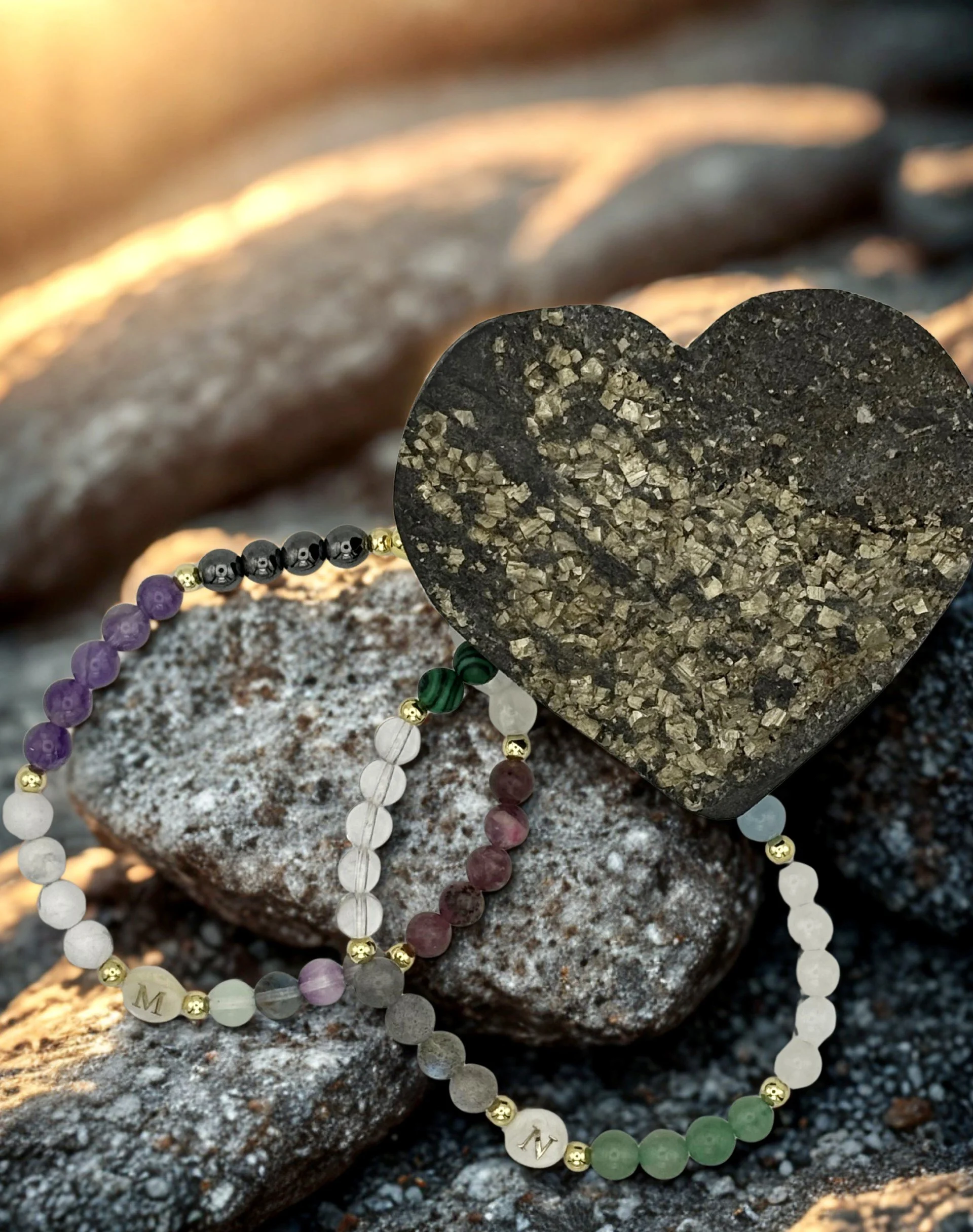 Close-up photo of heart-shaped stone with two crystal bracelets, placed on rocks with warm lighting.