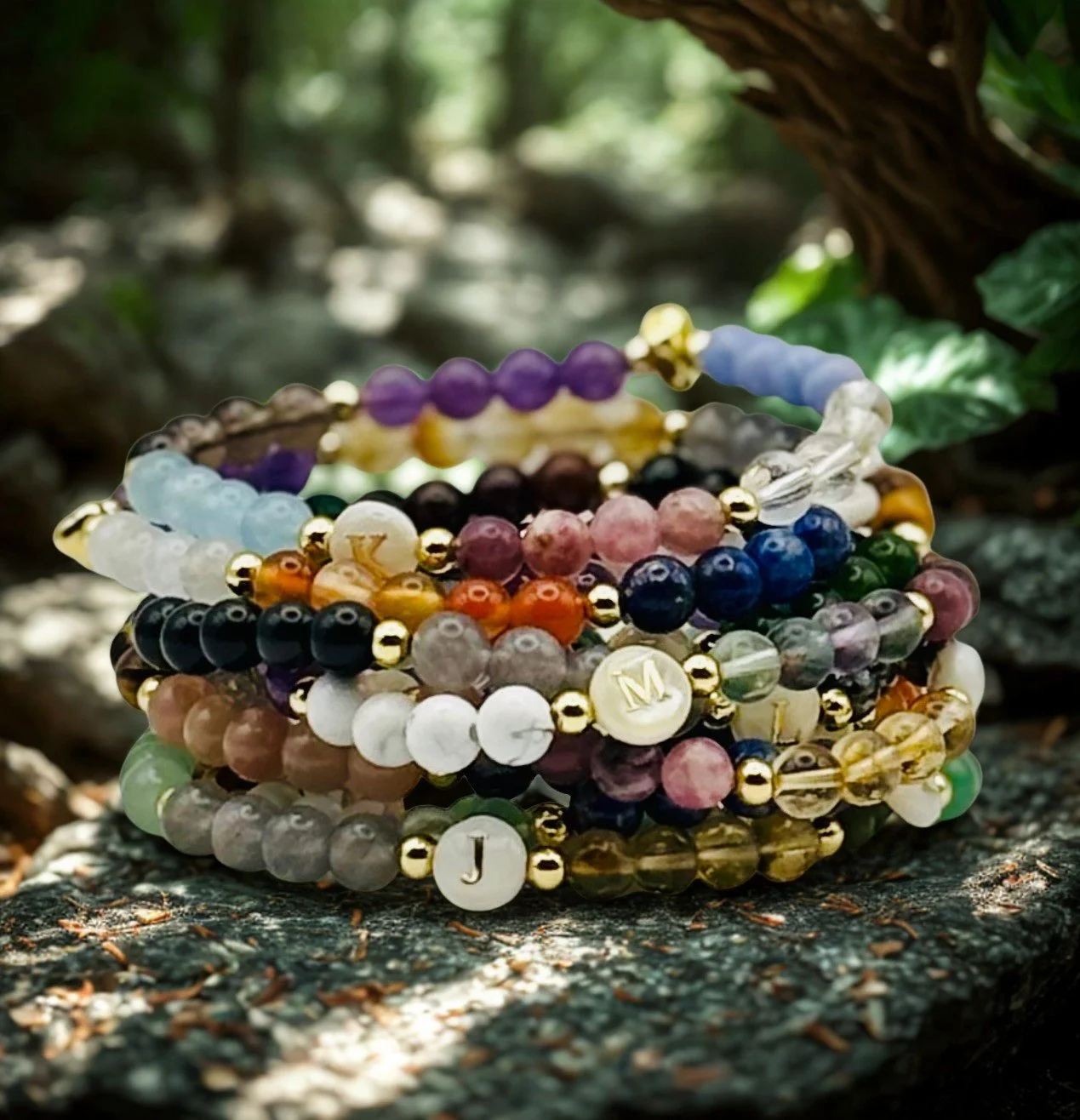 Multiple colourful crystals bracelets with gold accents, engraved letter charms, stacked on a rock outdoors with greenery in the background.