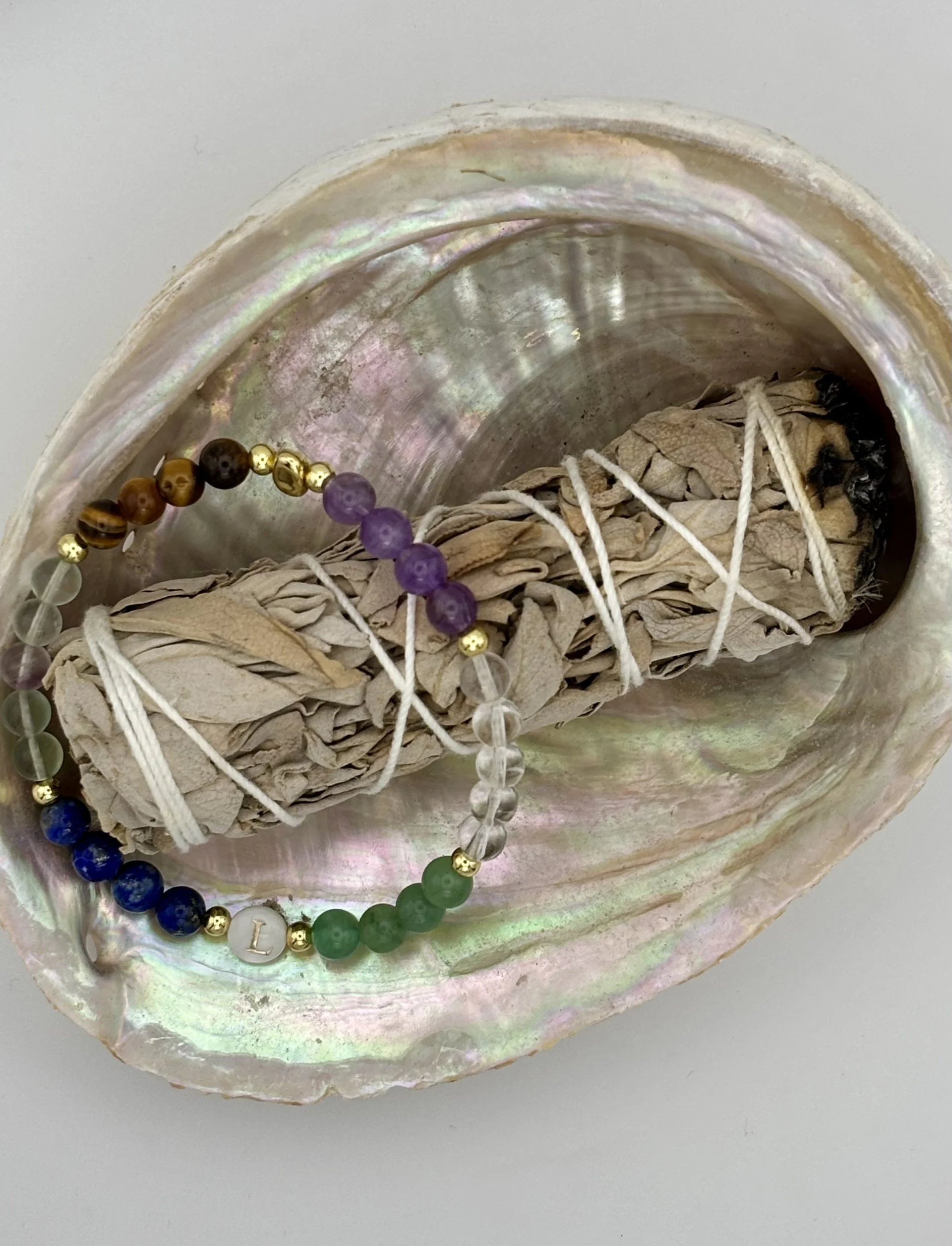 A large seashell with a slightly iridescent surface holding sage, and a crystal bracelet.