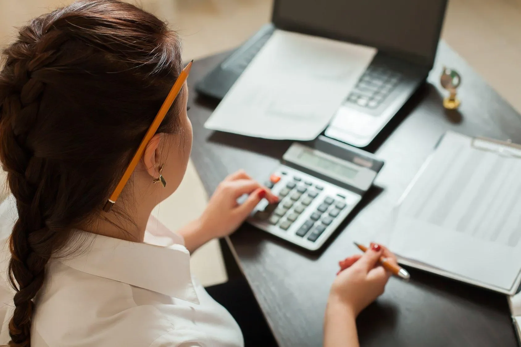 A woman working at a desk with a calculator, open notebook, printed documents, a laptop, and a key on the desk.