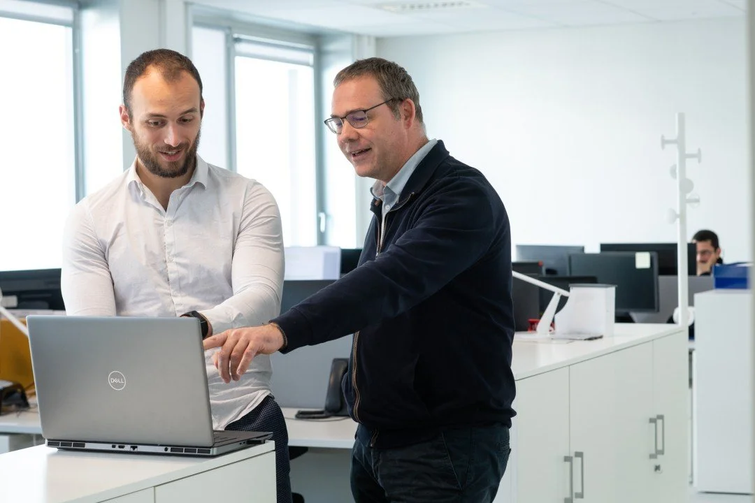 Two men discussing something on a laptop in an office environment.