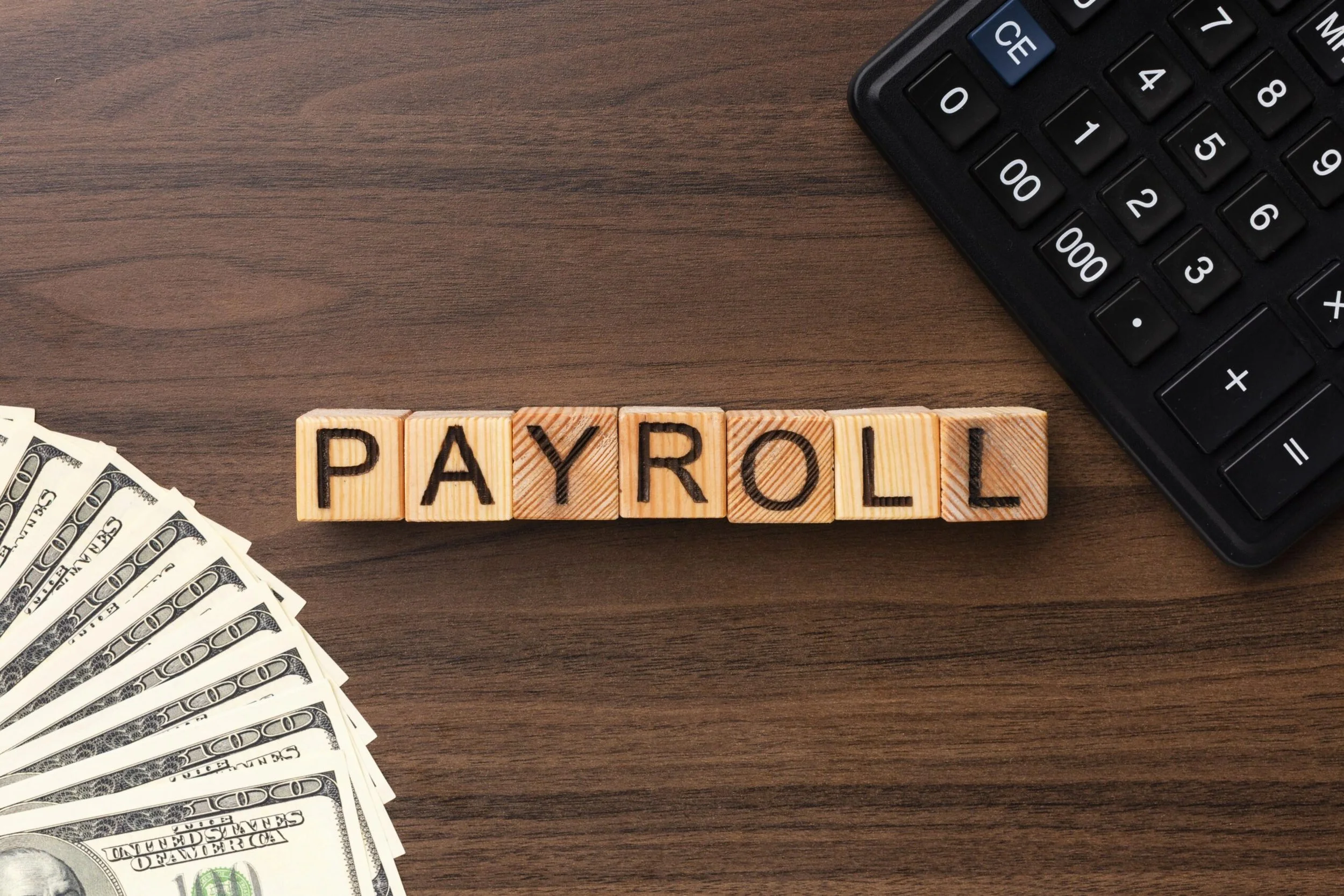 Wooden tiles spell out PAYROLL on a desk with US dollar bills, a calculator, and a wooden surface.