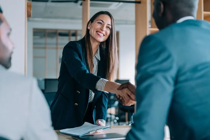 A woman smiling and shaking hands with a man in a business setting, while another man looks on.