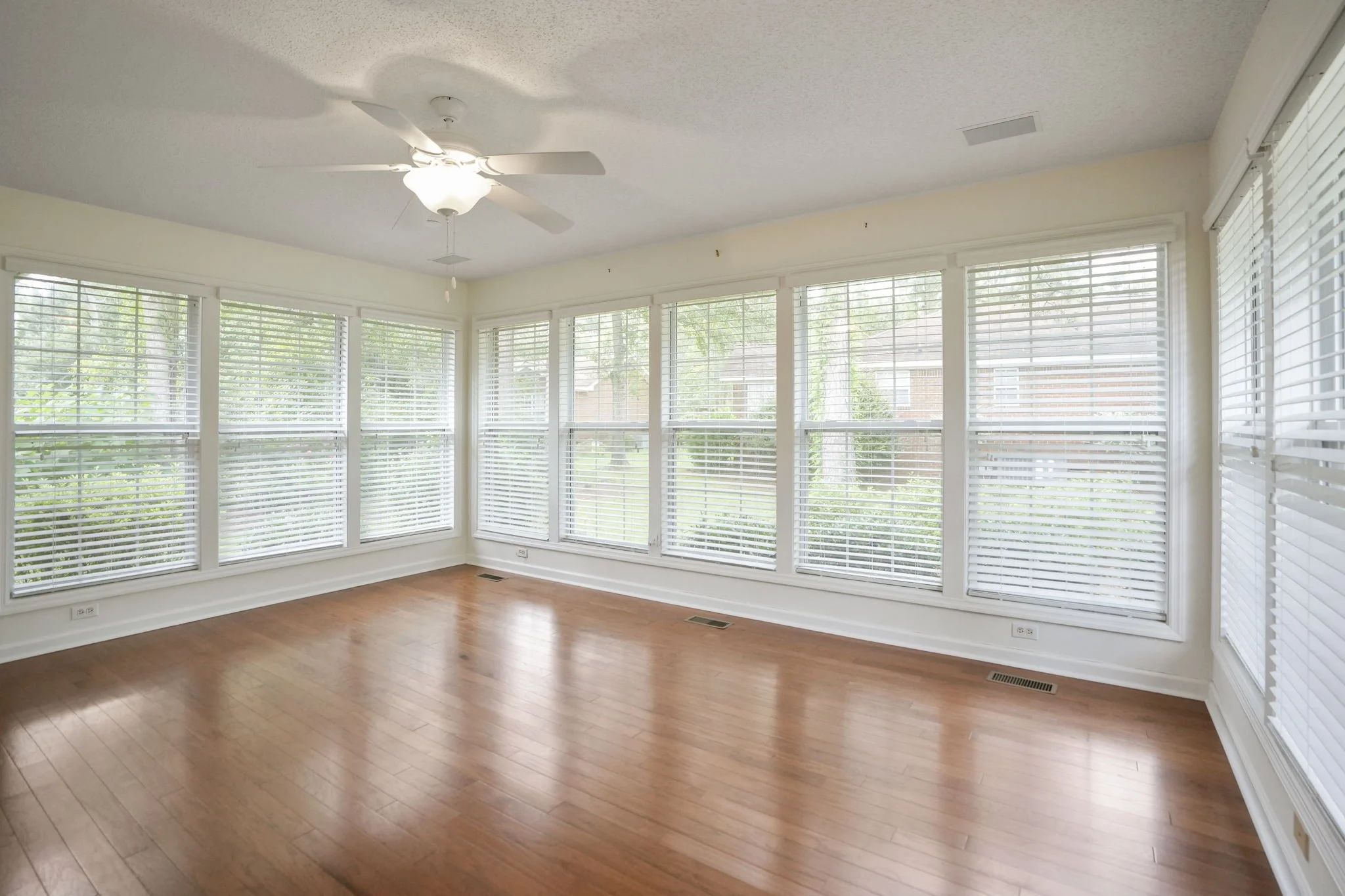 Empty room with large windows, wooden floor, white blinds, ceiling fan, and a view of a backyard with trees and nearby houses.