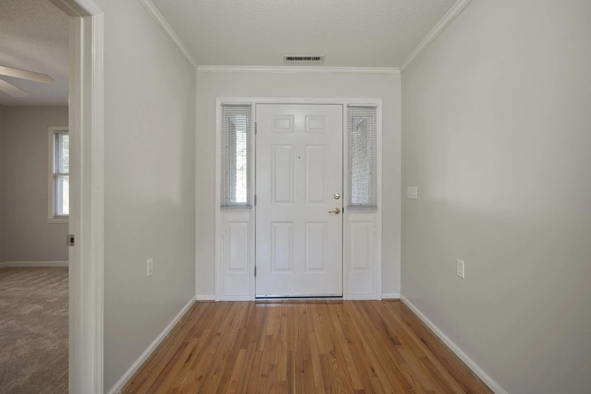 Empty foyer with a white door, two side windows with blinds, light-colored hardwood floors, and light-colored walls.