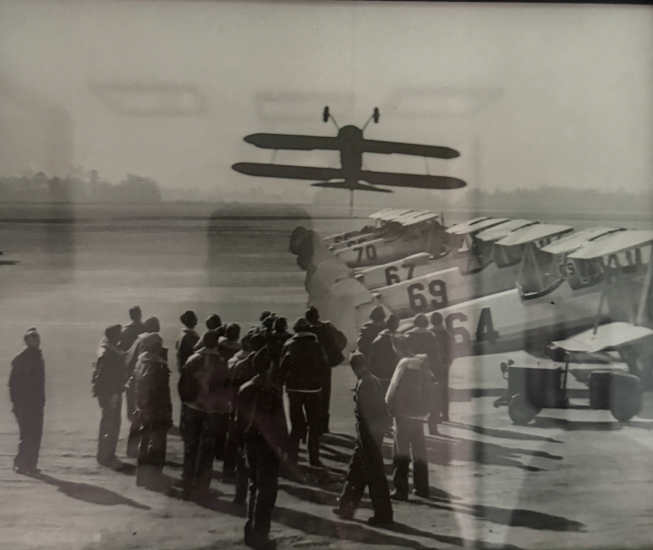 Black and white historical photo of an airplane with a biplane silhouette on the tail, parked on an airfield with a group of people gathered nearby, possibly during an aviation event or at an airport.