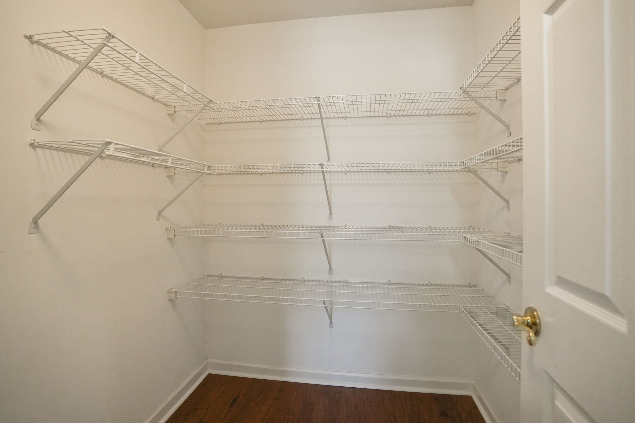 Empty white wire shelving unit in a walk-in closet with beige walls and a hardwood floor.