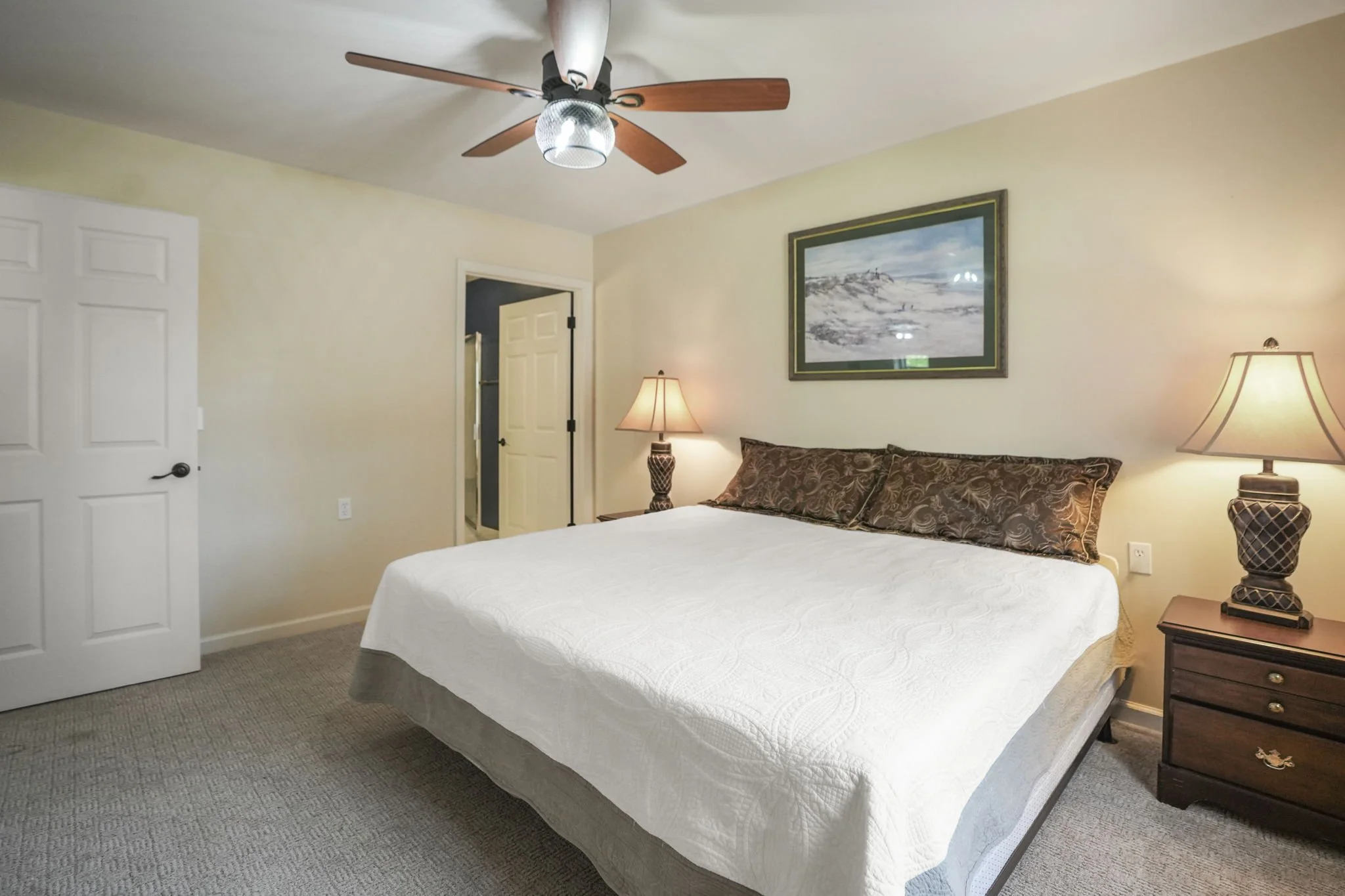 A bedroom with a large bed covered in a white quilt, two decorative pillows, a wooden nightstand with a lamp, a framed landscape painting on the wall, a ceiling fan with lights, and an open door leading to a bathroom.