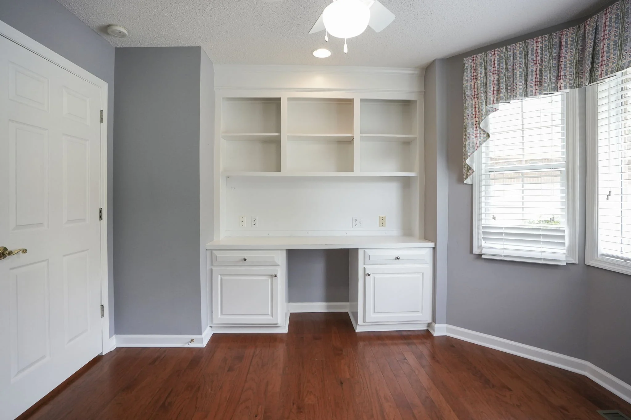 Empty room with gray walls, a built-in white desk with cabinets, open shelves above, brown hardwood flooring, and two windows with white blinds and a colorful valance.