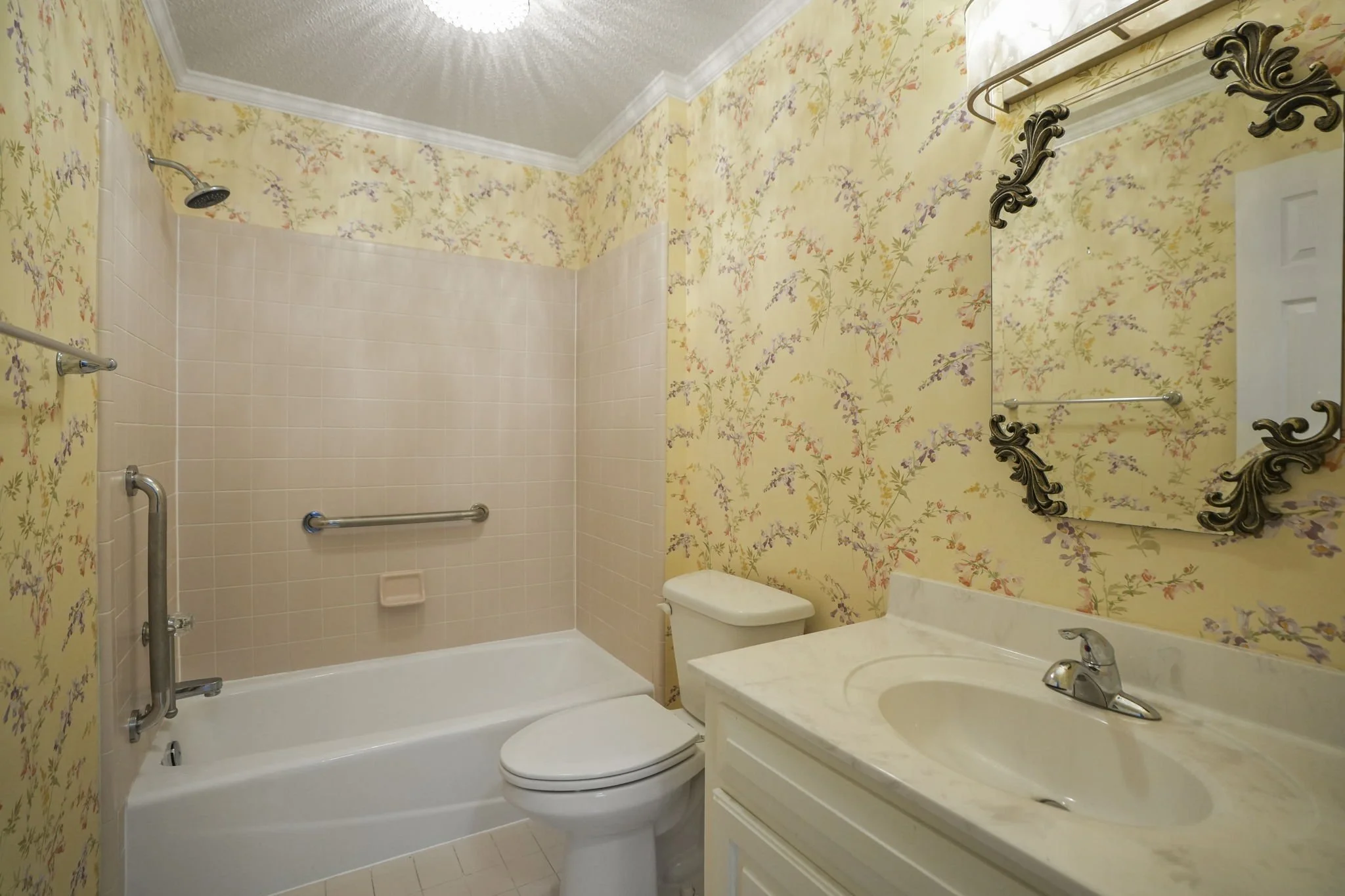 Bathroom with a bathtub, toilet, and vanity with mirror, yellow floral wallpaper, and a showerhead in the corner.