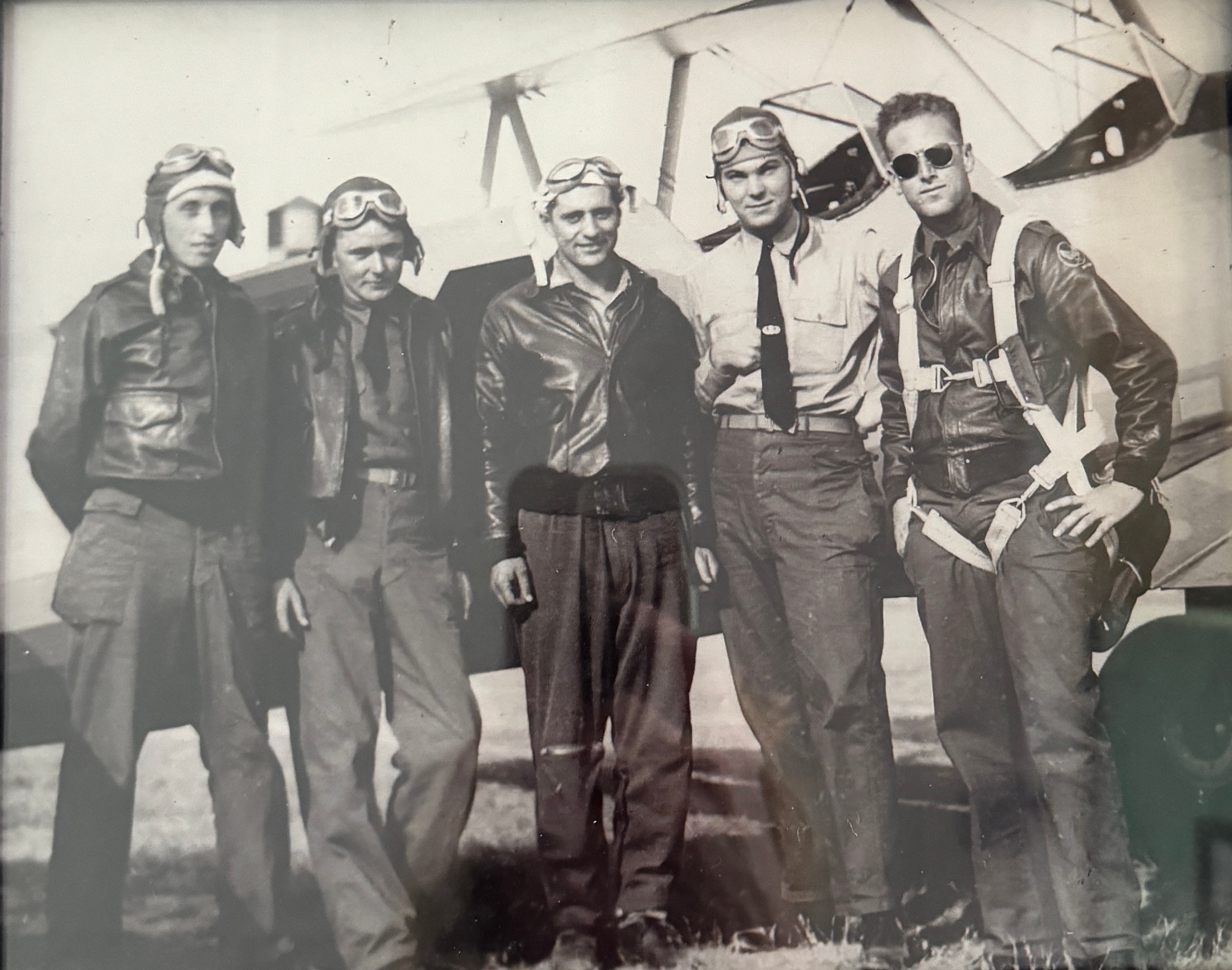 Black and white photograph of five individuals standing in front of an airplane, dressed in vintage flight gear including leather jackets, goggles, and parachutes, suggesting a historical aviation setting.