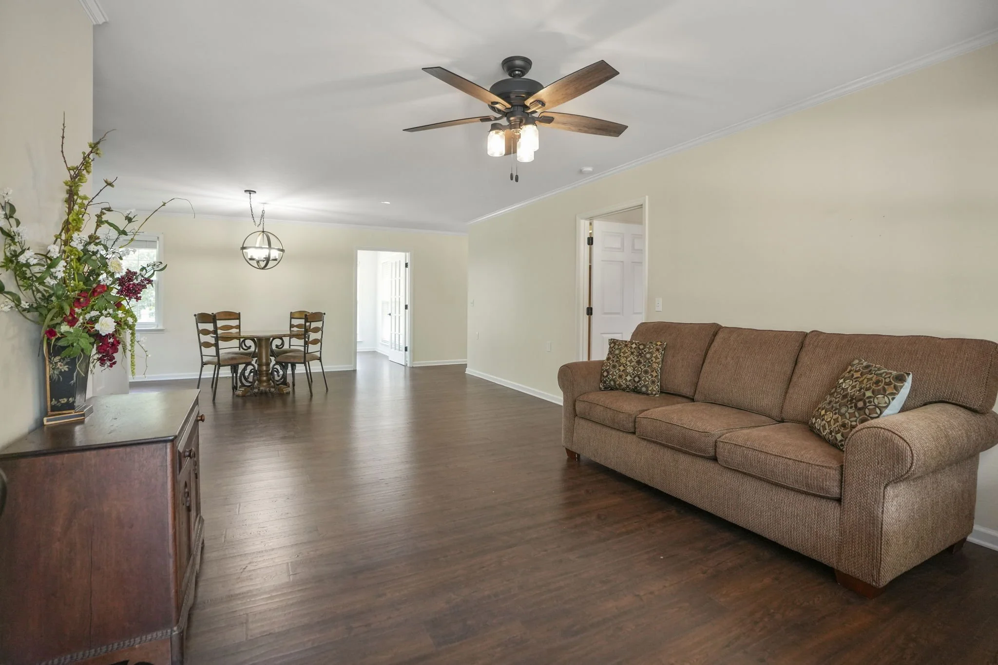 Living room with a brown couch, wooden flooring, a ceiling fan, and a dining area in the background with a round table and four chairs.