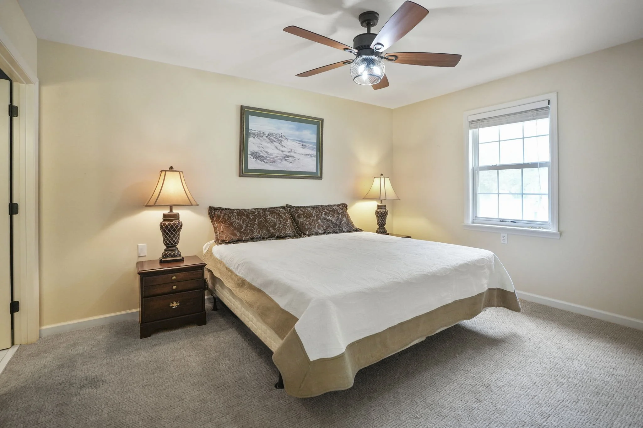 A tidy bedroom with a bed, two nightstands with lamps, a window, a ceiling fan, and a landscape painting on the wall.