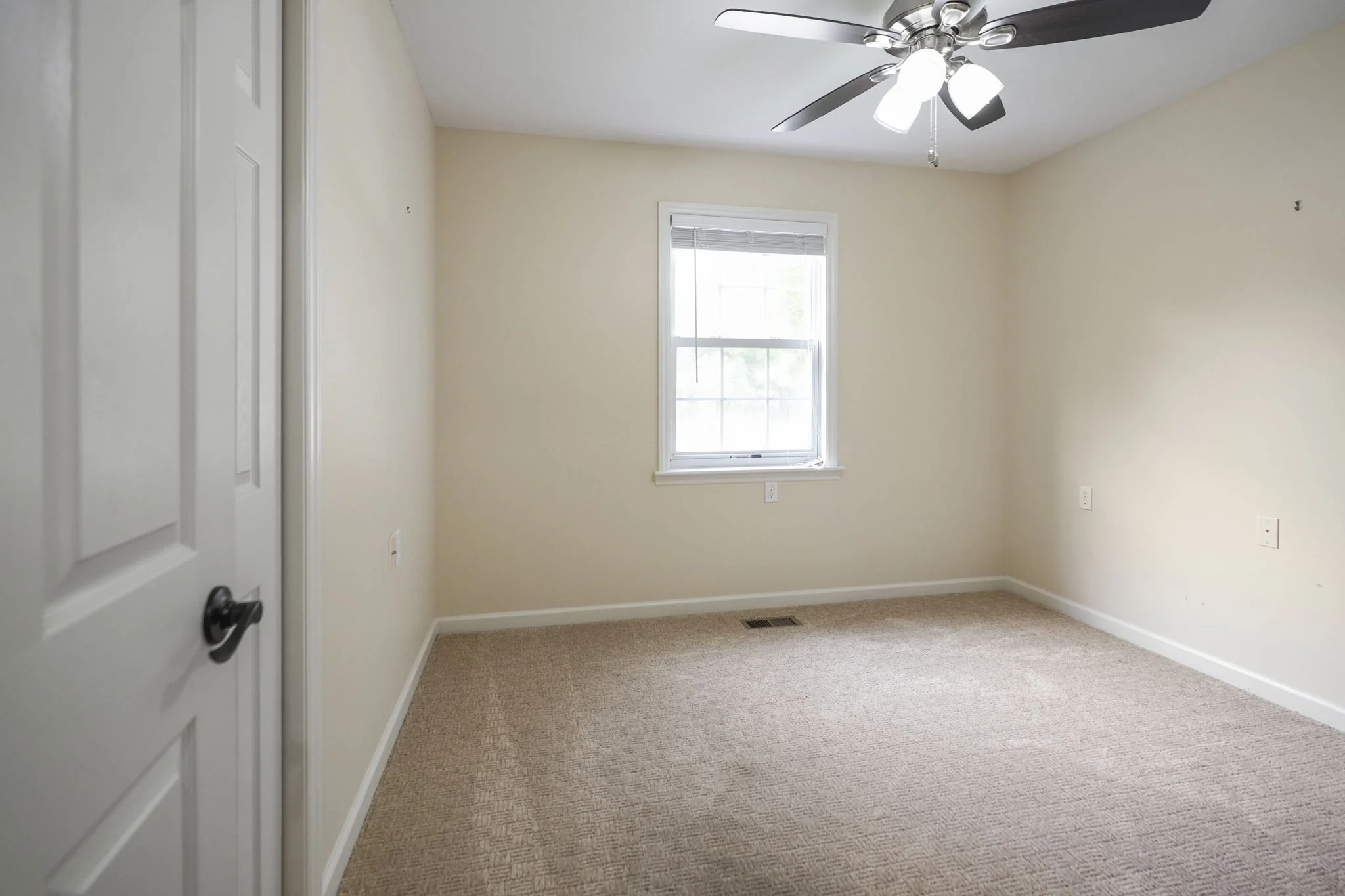 Empty beige carpeted room with a window, ceiling fan with lights, and light-colored walls.