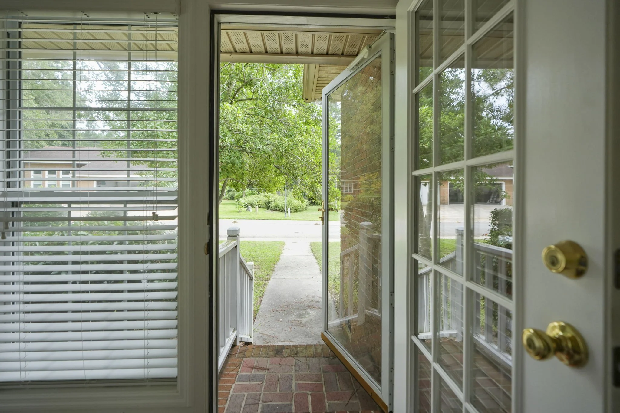 View from inside a house looking out through a screening door onto a porch and a quiet suburban street with grass and trees.