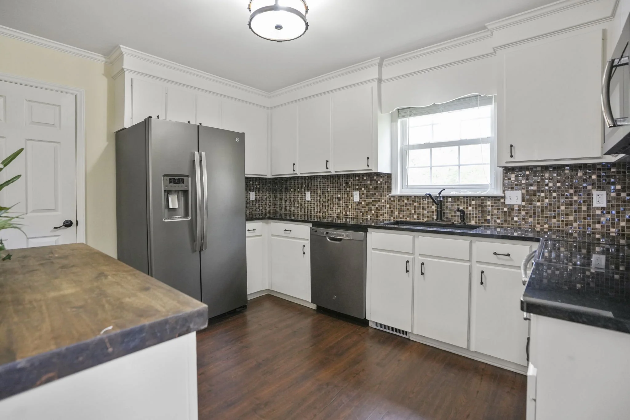 Modern kitchen with white cabinets, black countertops, brown mosaic tile backsplash, stainless steel refrigerator, dishwasher, and microwave, hardwood floors, window above sink.