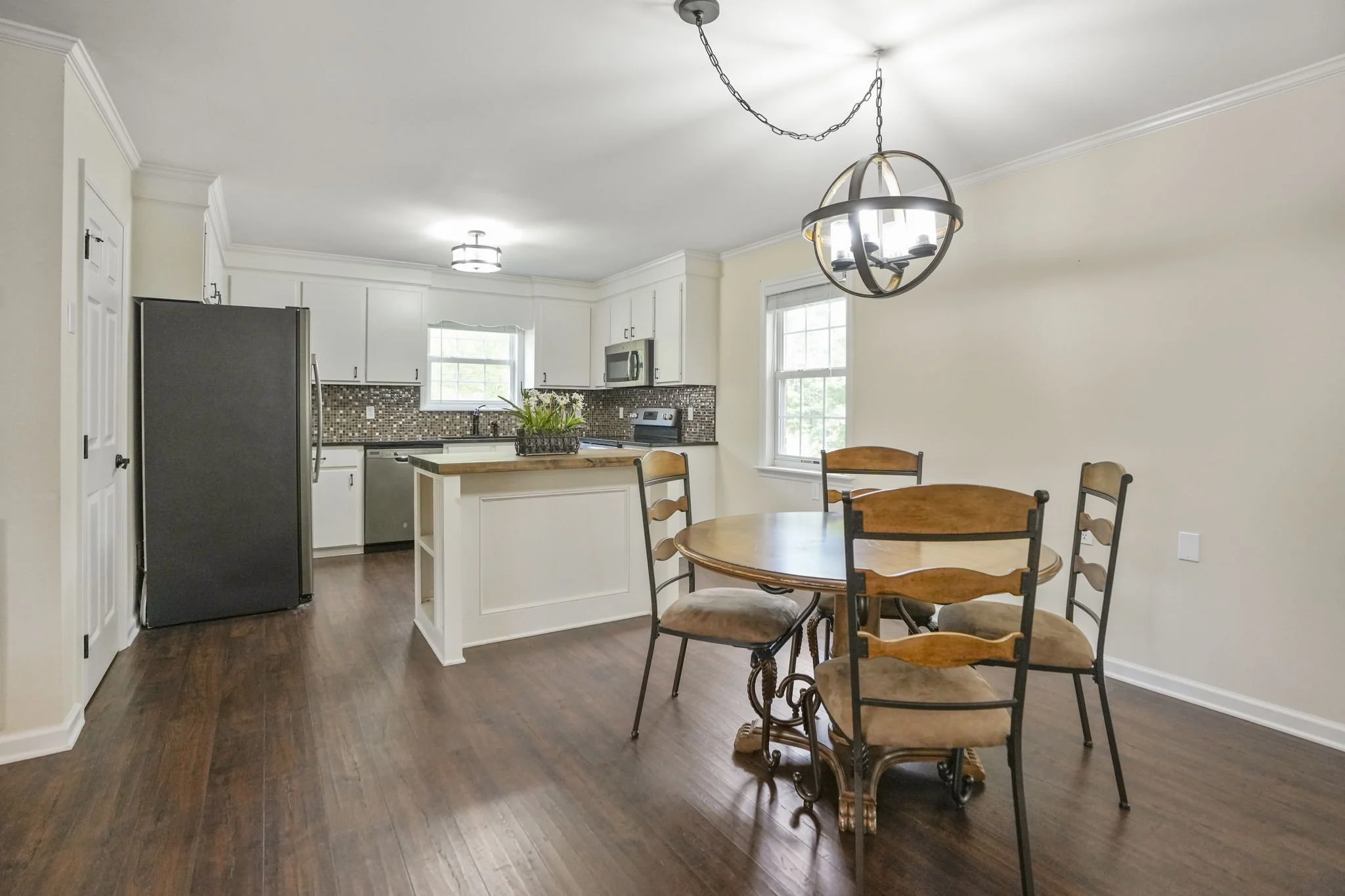 Interior view of a kitchen and dining area with hardwood floors, white cabinets, a mosaic tile backsplash, stainless steel appliances, and a round wooden dining table with four chairs, illuminated by a chandelier.