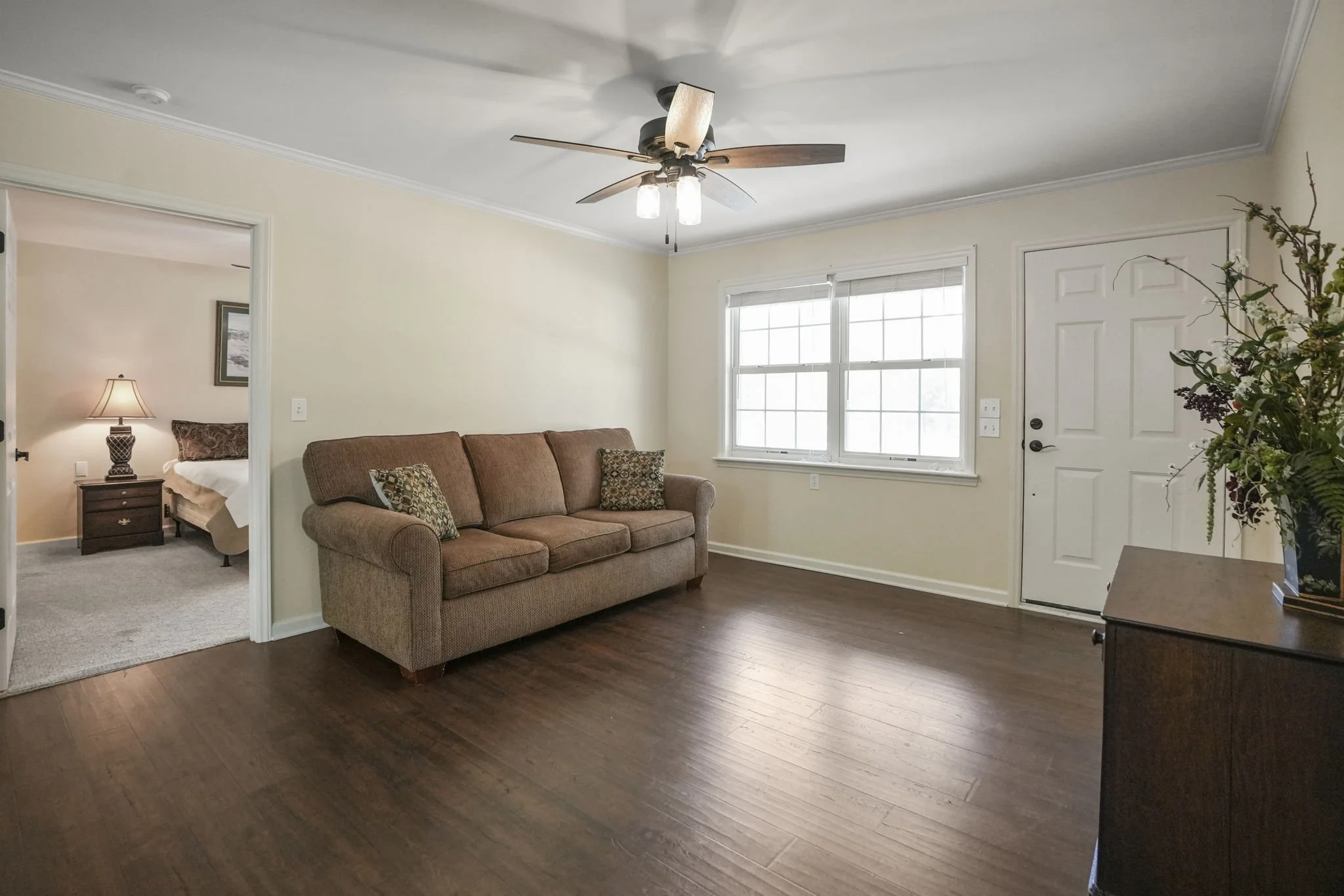 Living room with brown sofa, wooden floor, large window, ceiling fan, and adjacent bedroom with lamp and bed.