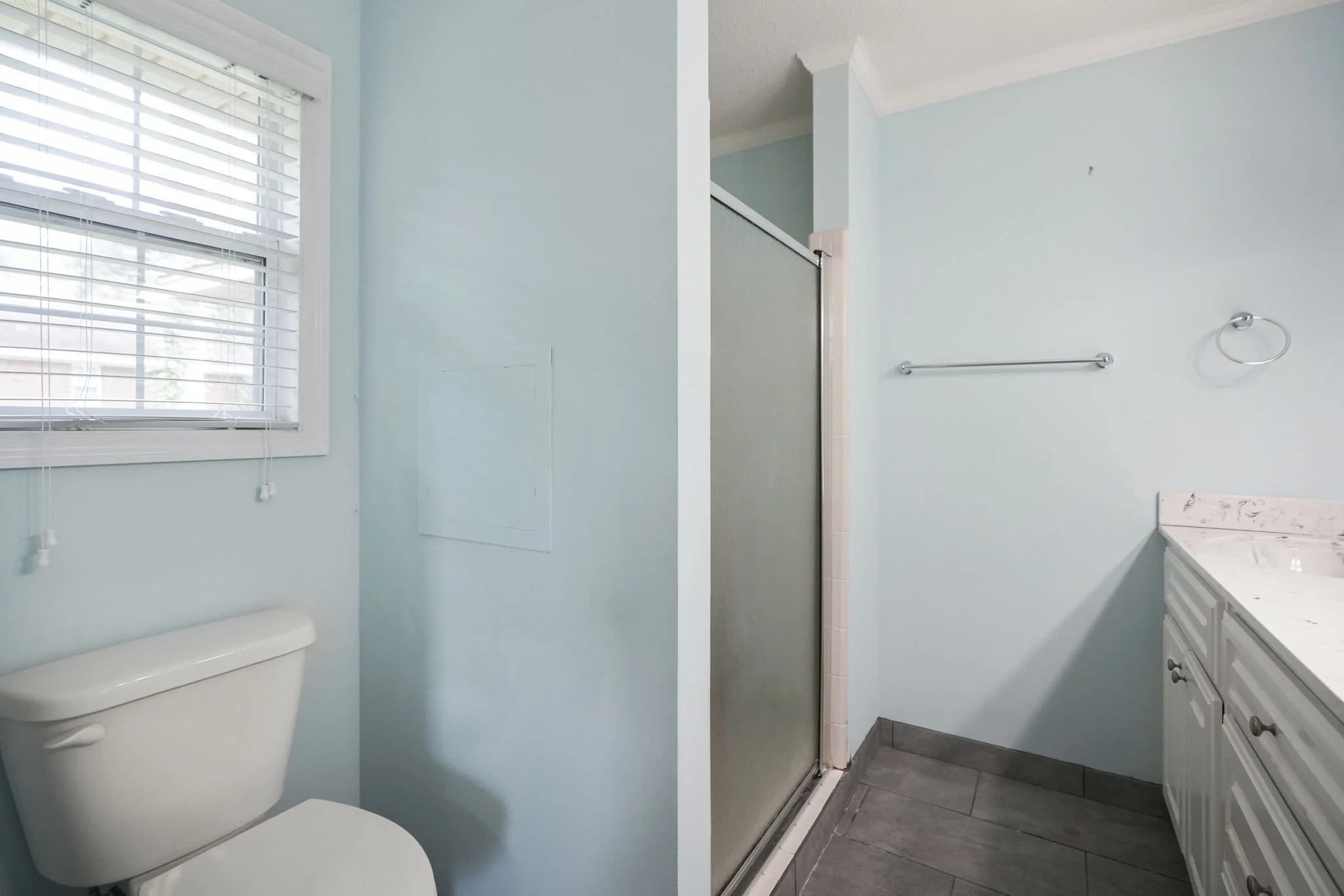 Bathroom with light blue walls, white toilet, window with blinds, shower with frosted glass door, and white vanity with marble countertop.